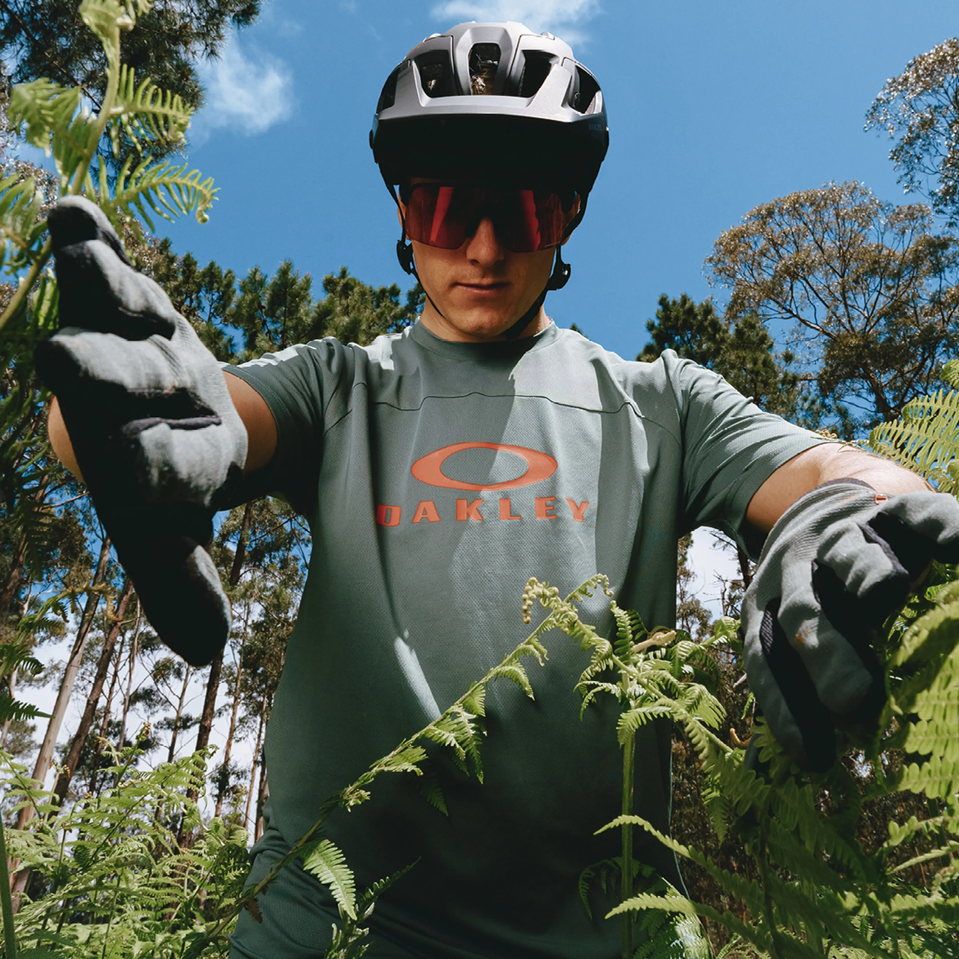 A cyclist wearing a helmet, sunglasses, and gloves stands among green ferns, against a backdrop of trees and a clear blue sky.