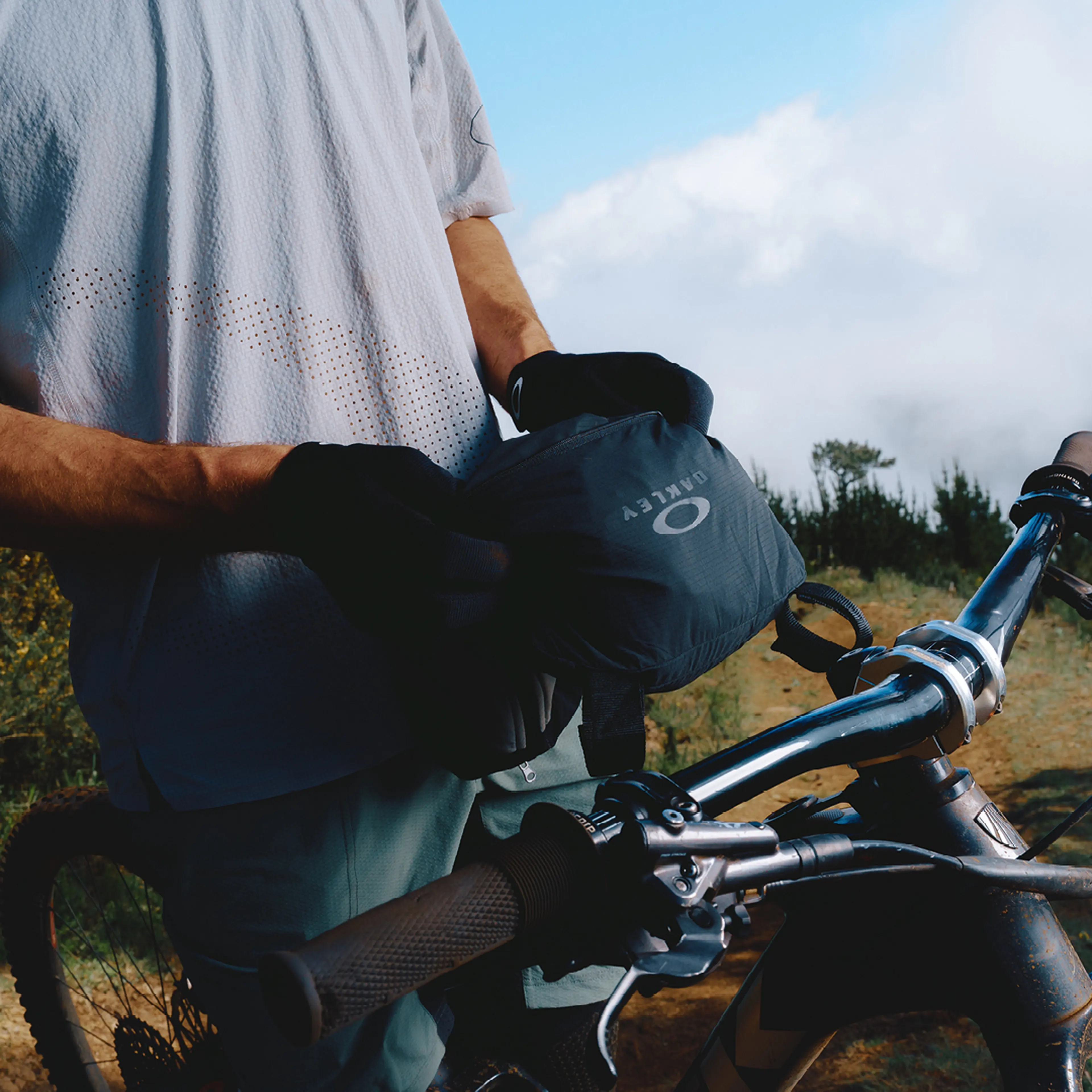 A person with gloves holds a black Oakley bag near a mountain bike on a dirt trail with cloudy sky in the background.