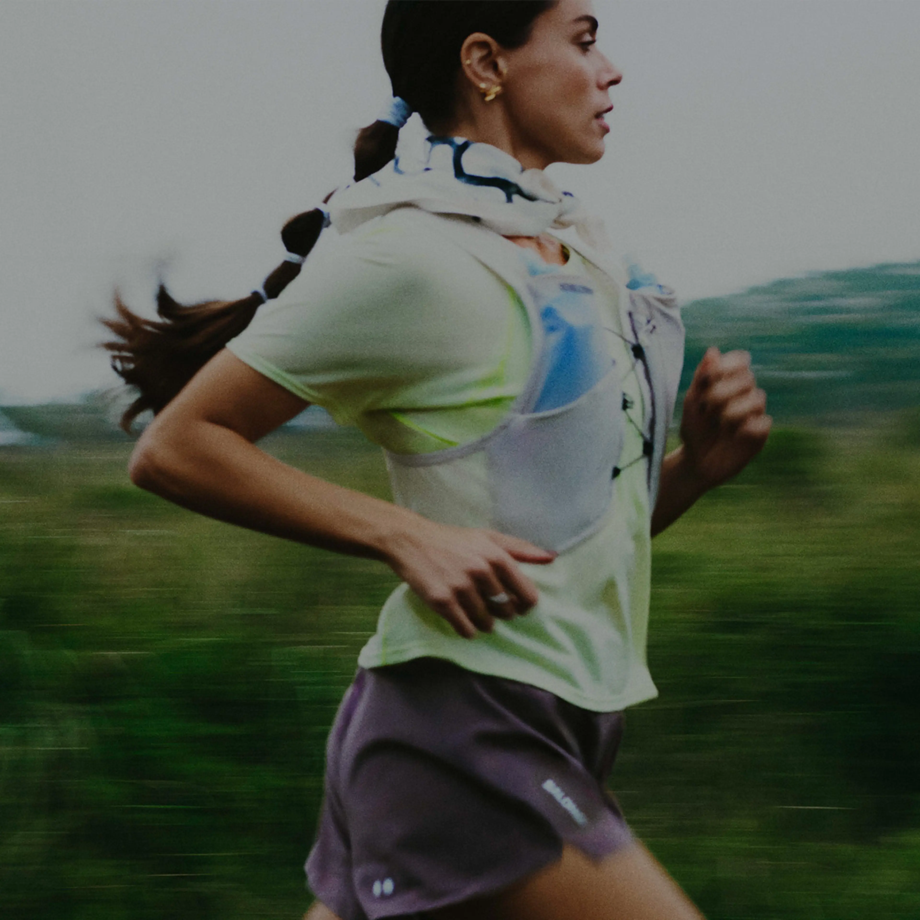 A woman with a ponytail runs outdoors wearing a light shirt, shorts, and a hydration vest, with greenery blurred in the background.