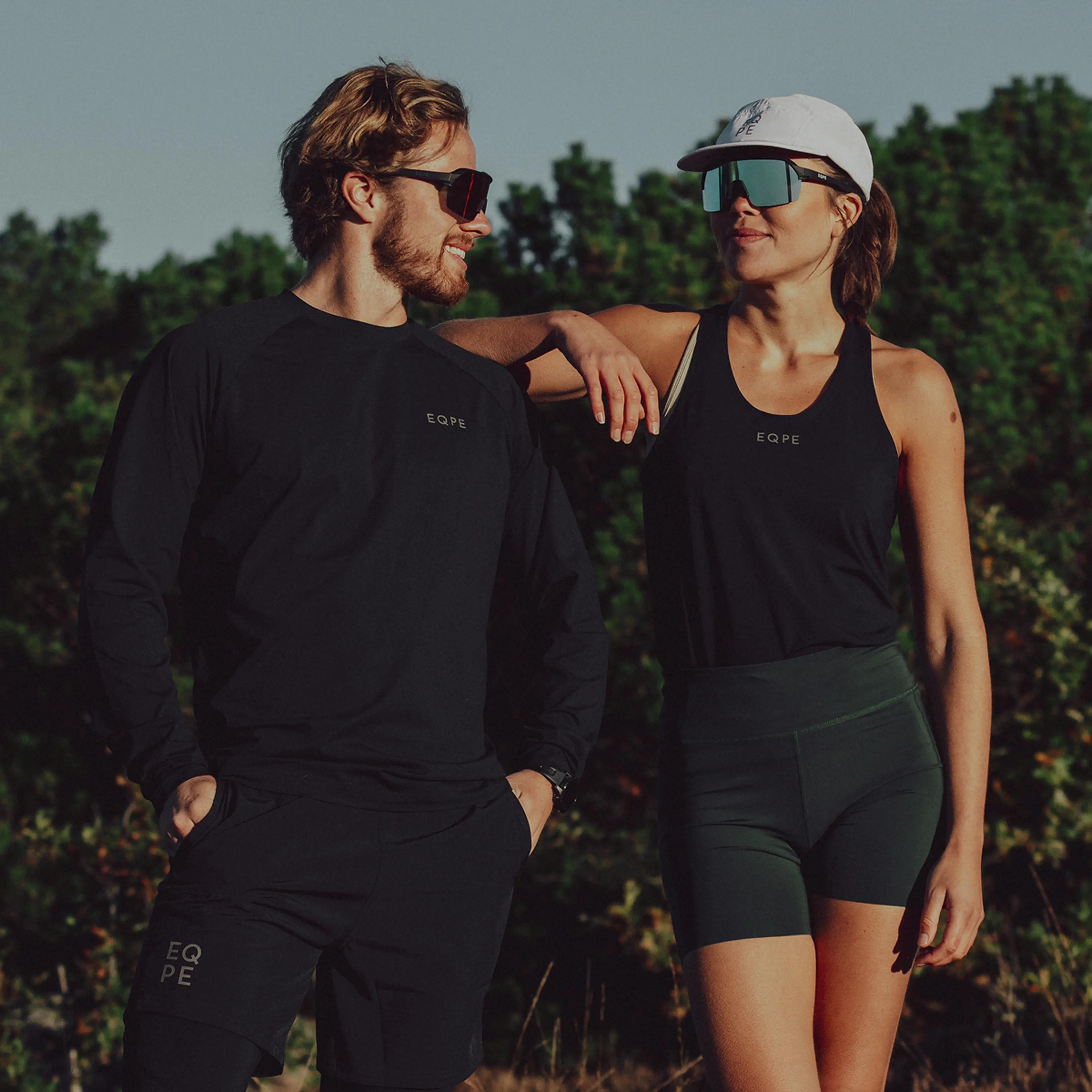 A man and a woman in athletic wear stand outdoors, both wearing sunglasses. The woman leans on the man's shoulder. Trees are in the background.
