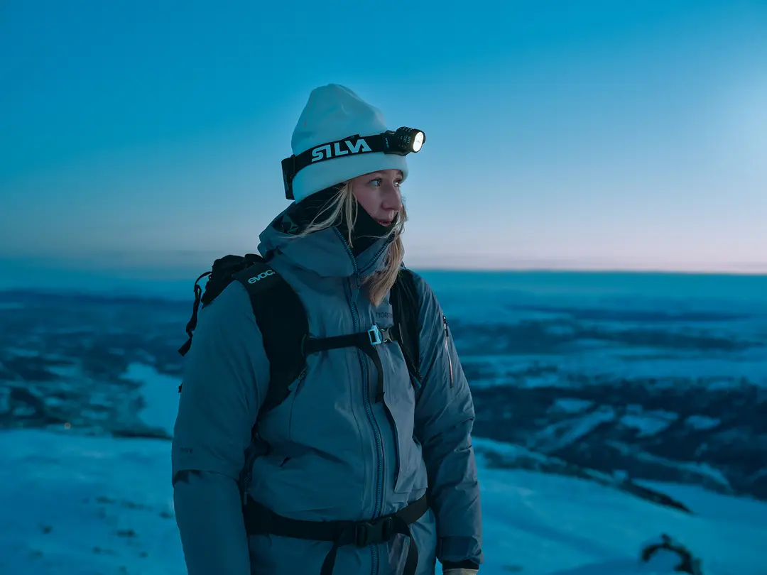 A person in winter gear with a headlamp stands on a snowy mountain at dusk, gazing into the distance against a blue-hued sky.
