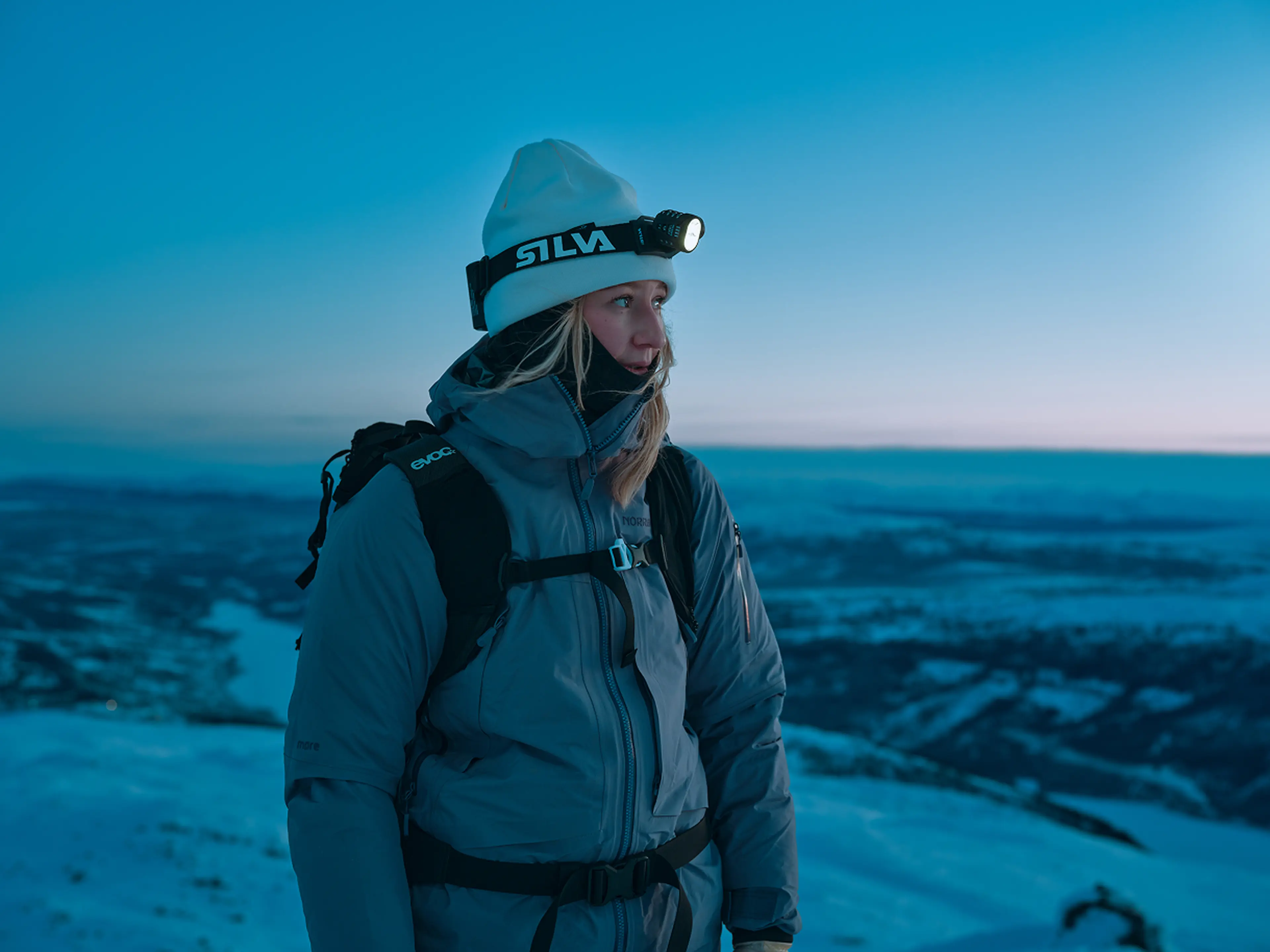 A person in winter gear with a headlamp stands on a snowy mountain at dusk, gazing into the distance against a blue-hued sky.