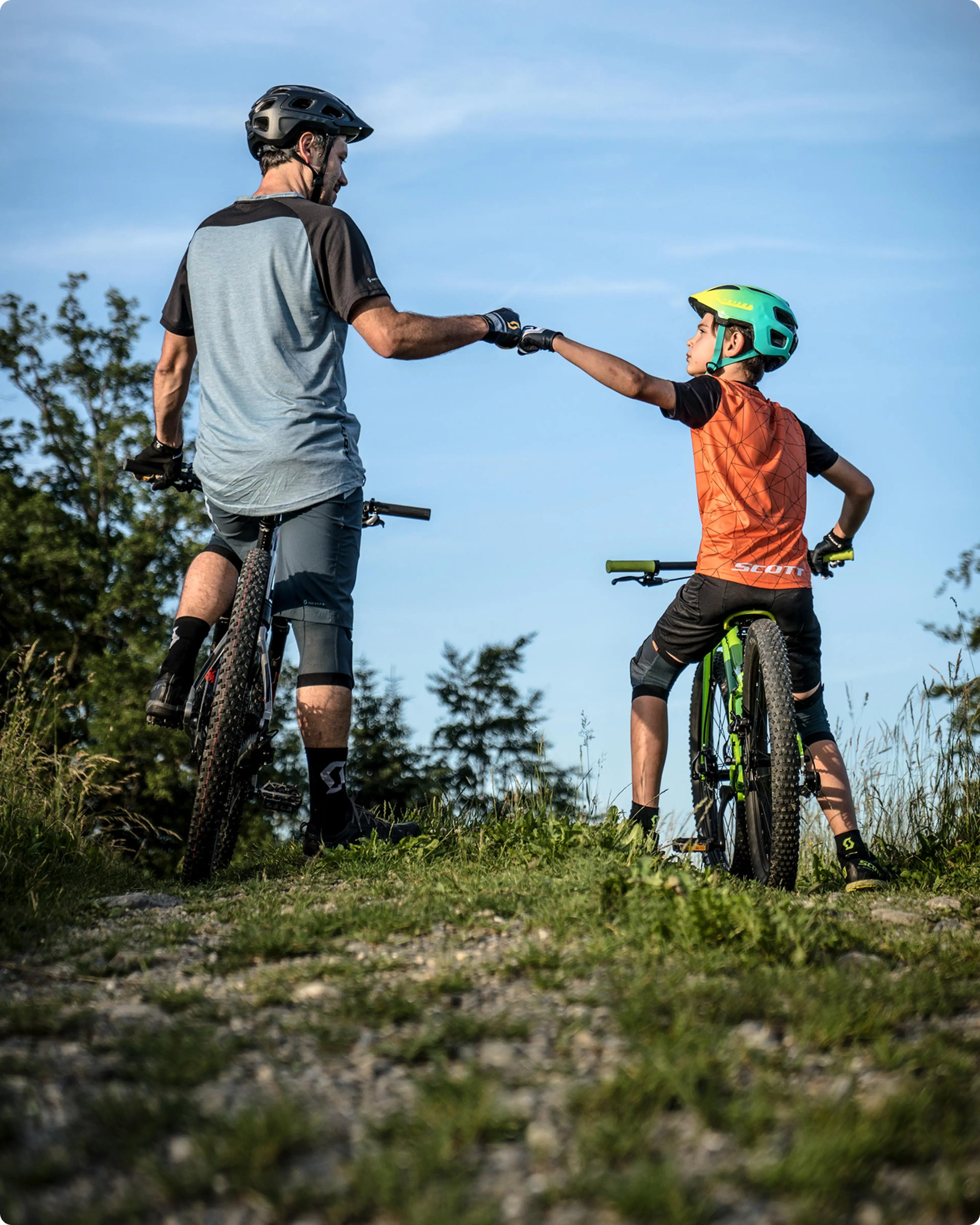Two cyclists on a trail, an adult and a child, give each other a fist bump while wearing helmets and casual biking attire.