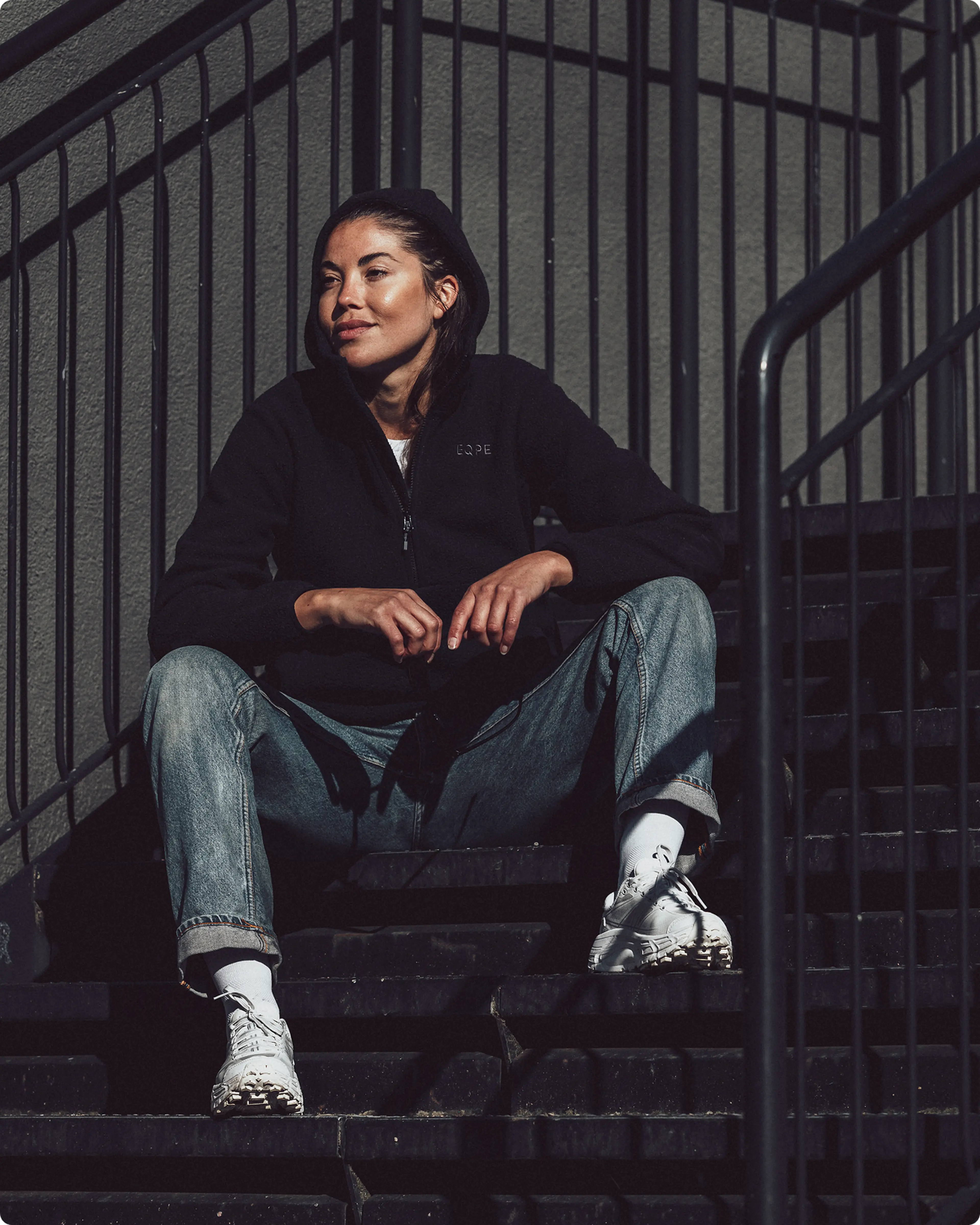 Person in a black hoodie and jeans sits on outdoor stairs, lit by sunlight, with metal railings in the background.