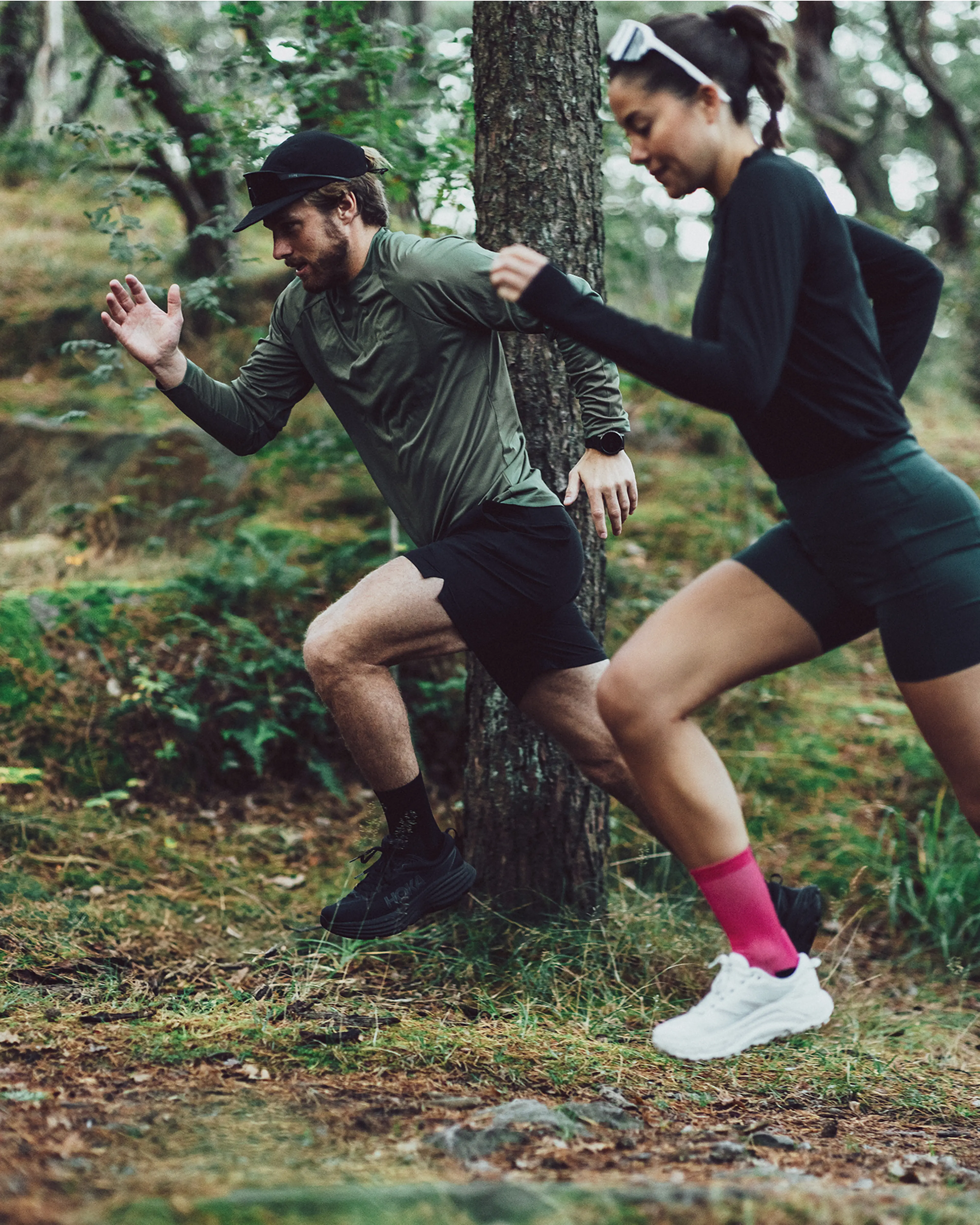 Two people trail running through a forest, wearing sporty outfits and focusing on their stride amidst the trees.