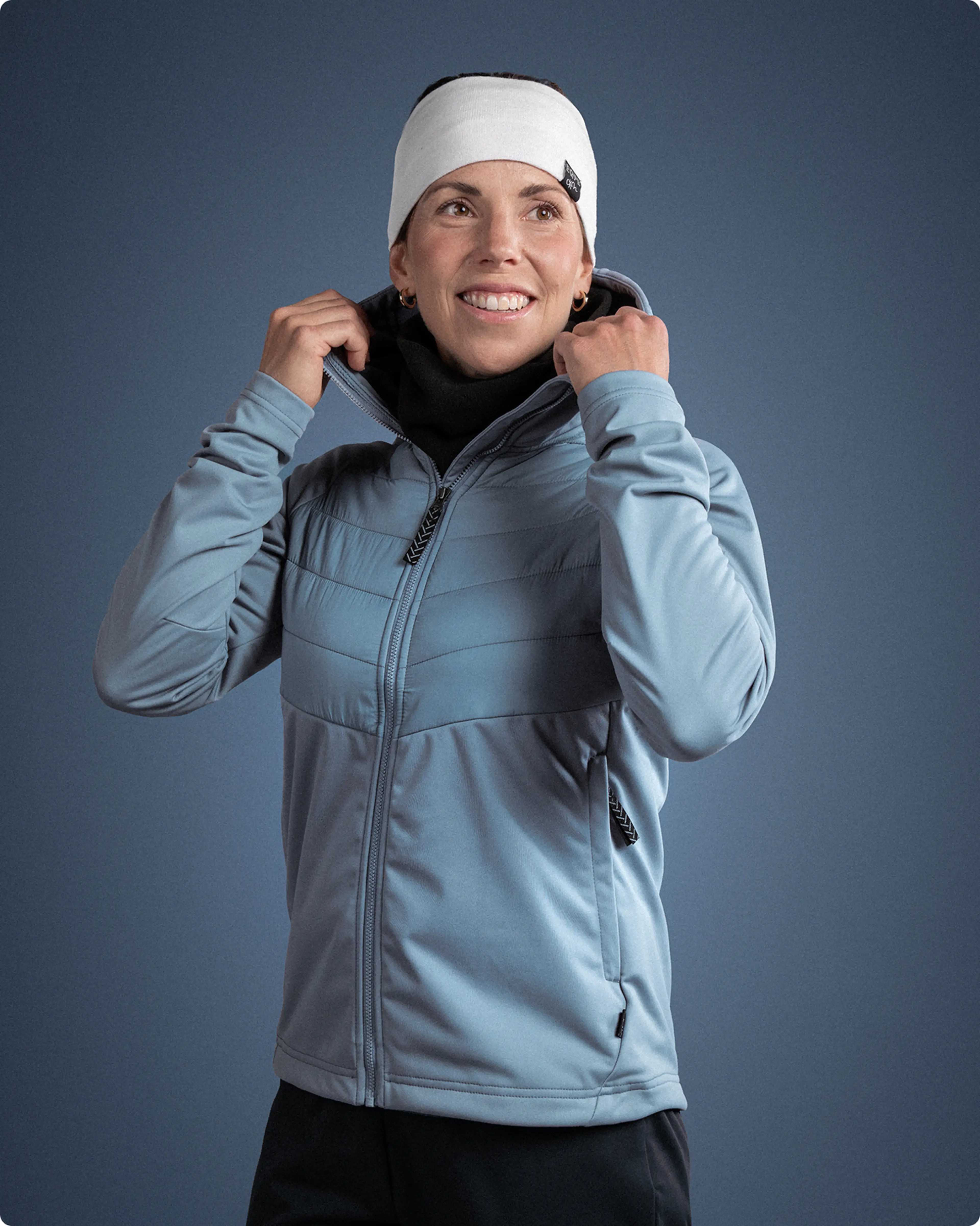 Person in a blue jacket and white headband smiling, adjusting their collar against a plain blue background.