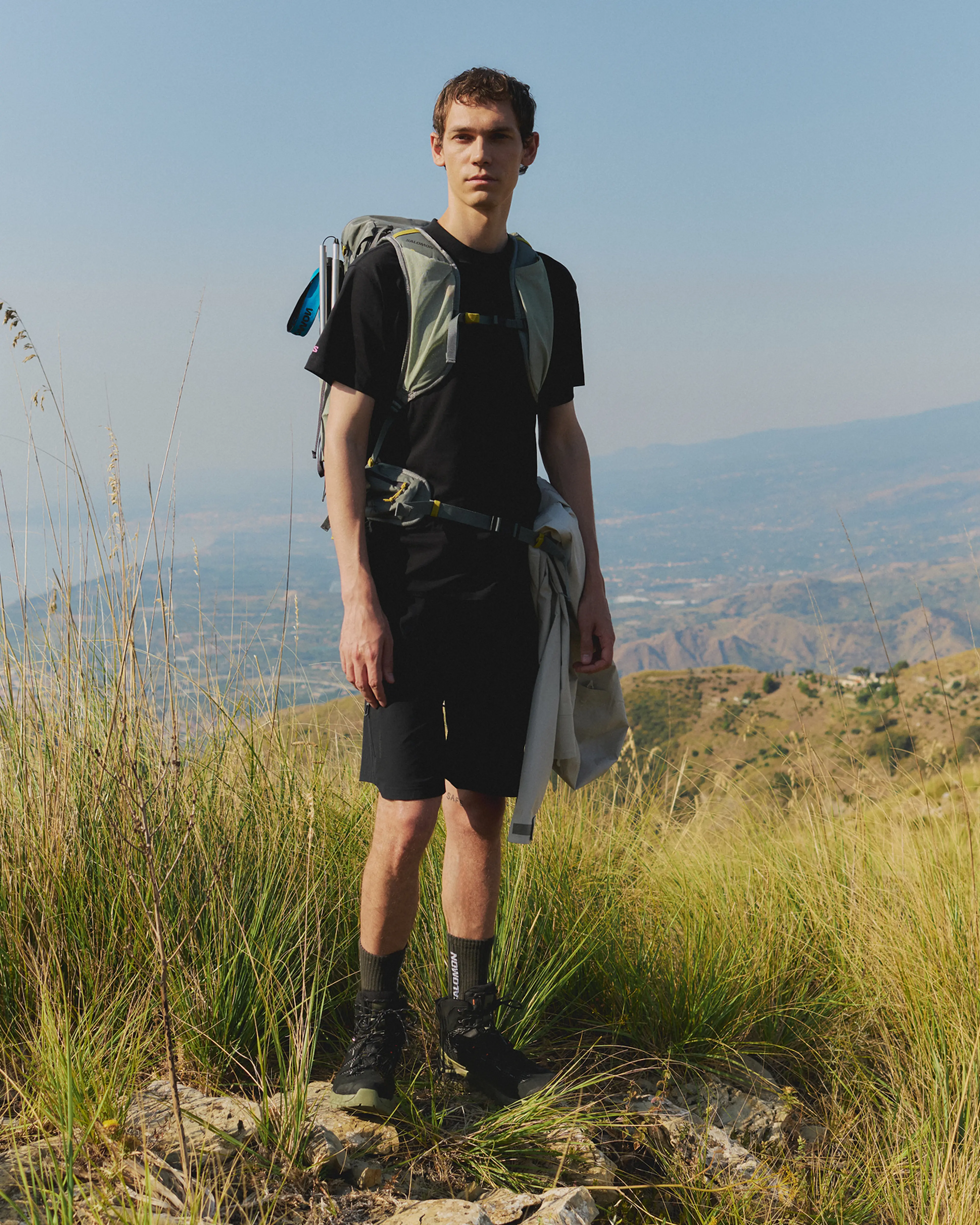 A person stands on a grassy hilltop wearing a backpack, black outfit, and hiking boots, with a scenic view of mountains in the background.