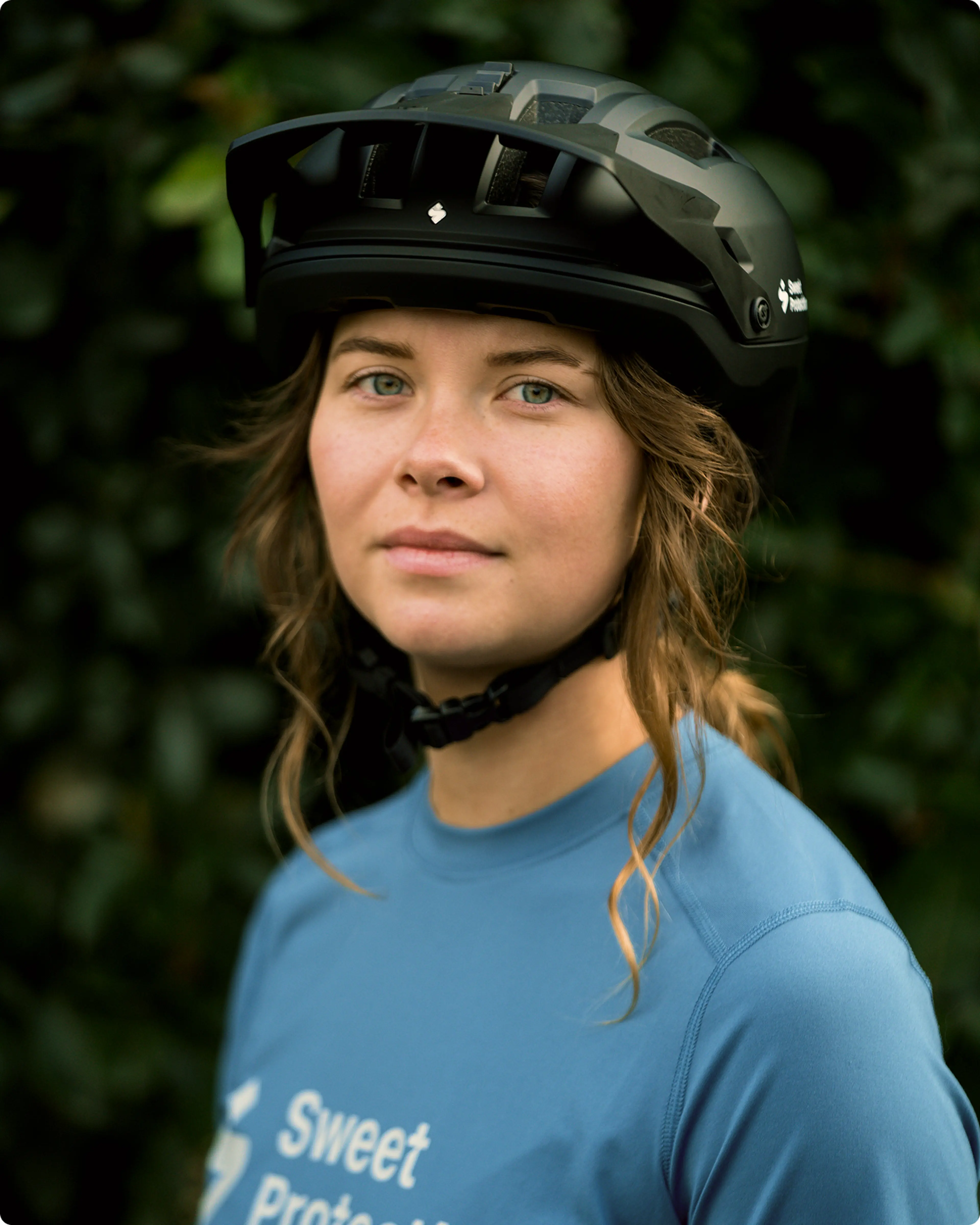 A person wearing a black bicycle helmet and a blue "Sweet Protection" shirt stands against a leafy background, looking confidently at the camera.
