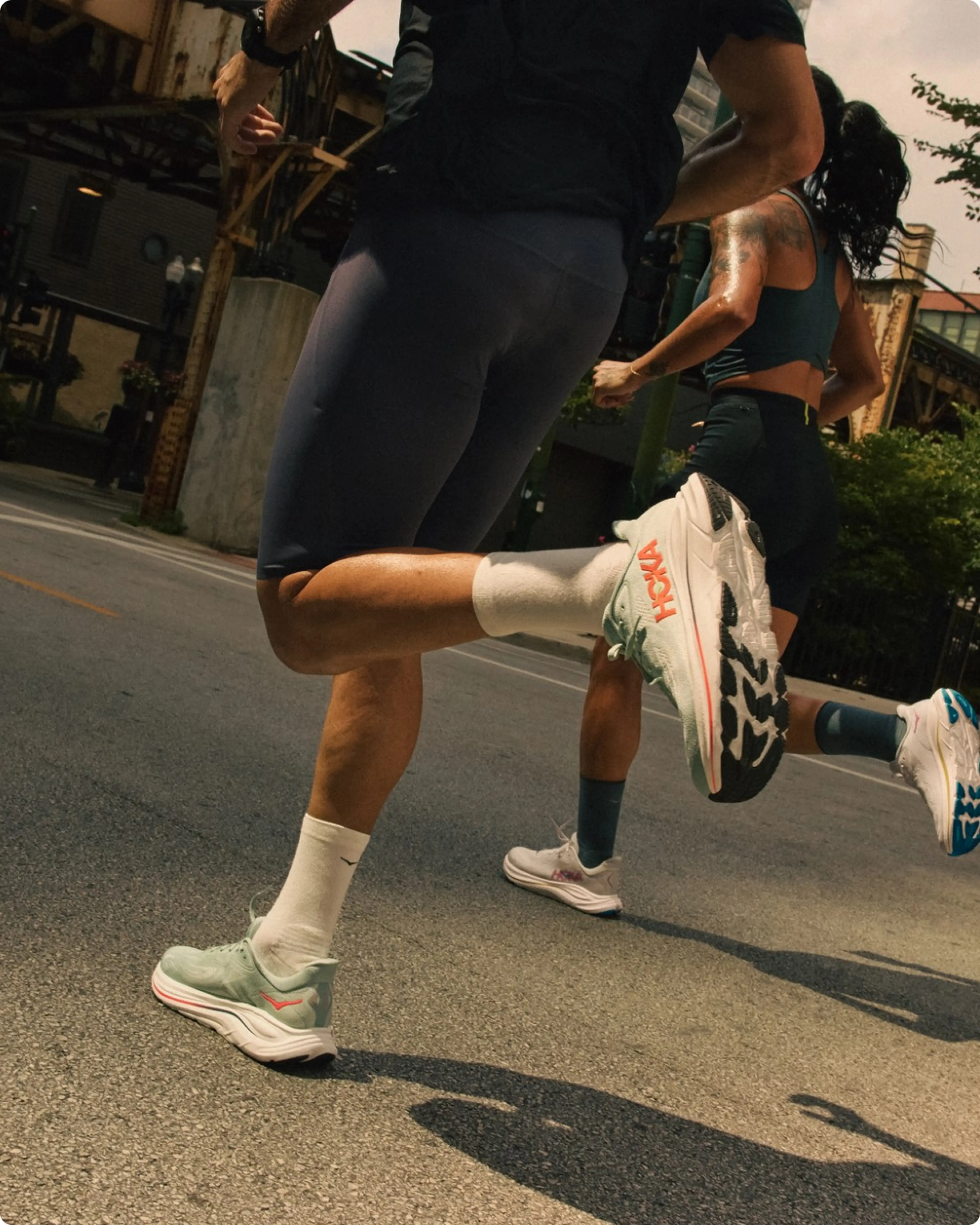 Two people running on a city street, focusing on their lower legs and colorful athletic shoes, with urban buildings in the background.