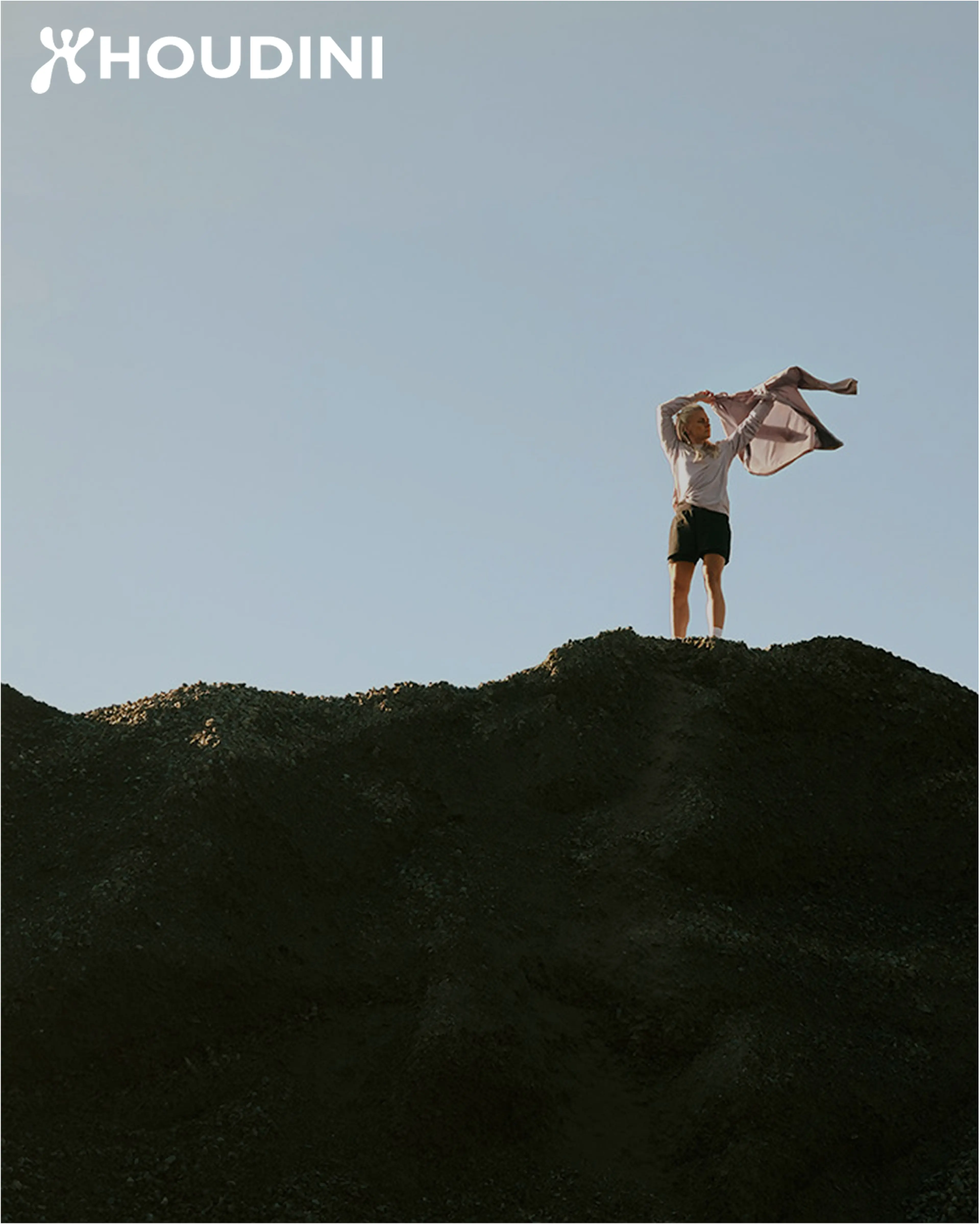 Person standing atop a large hill, holding a jacket against a clear sky. Houdini logo in the top left corner.