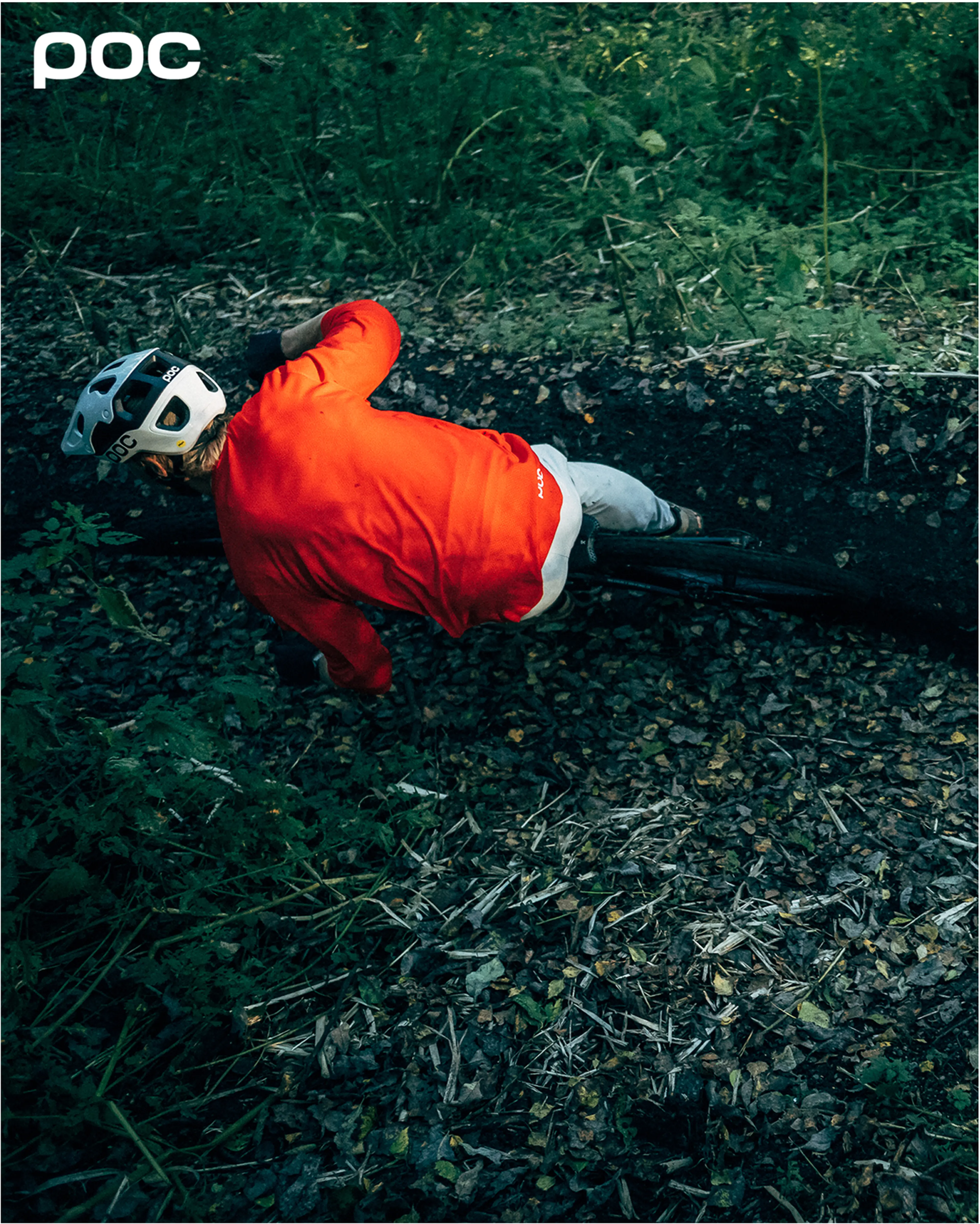 A cyclist in a red jacket and helmet rides through a forest trail, leaning into a turn on a dirt path surrounded by greenery.  With Poc branding in the corner.