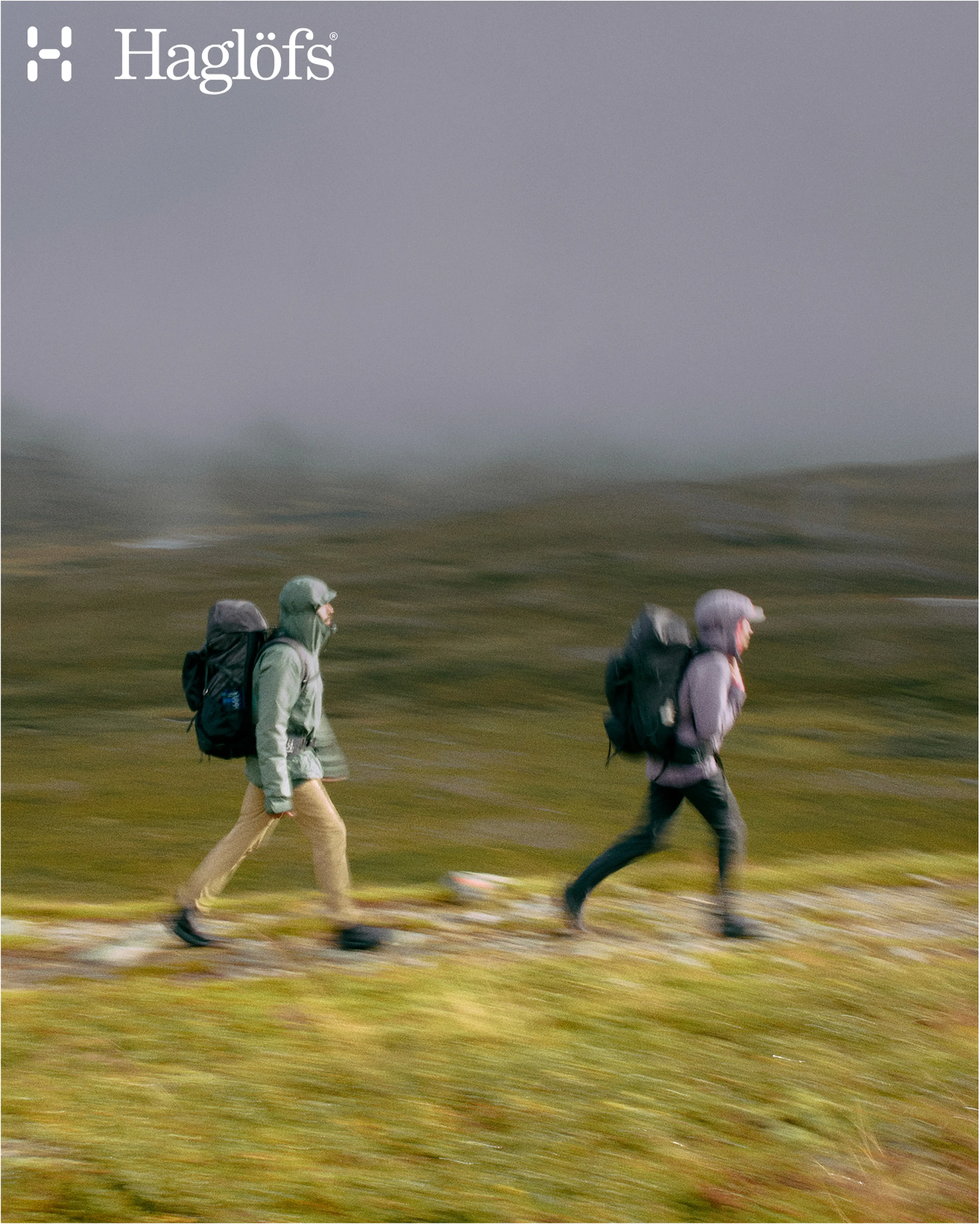 Two hikers wearing jackets and backpacks walk briskly through a foggy landscape, featuring the Haglöfs logo.