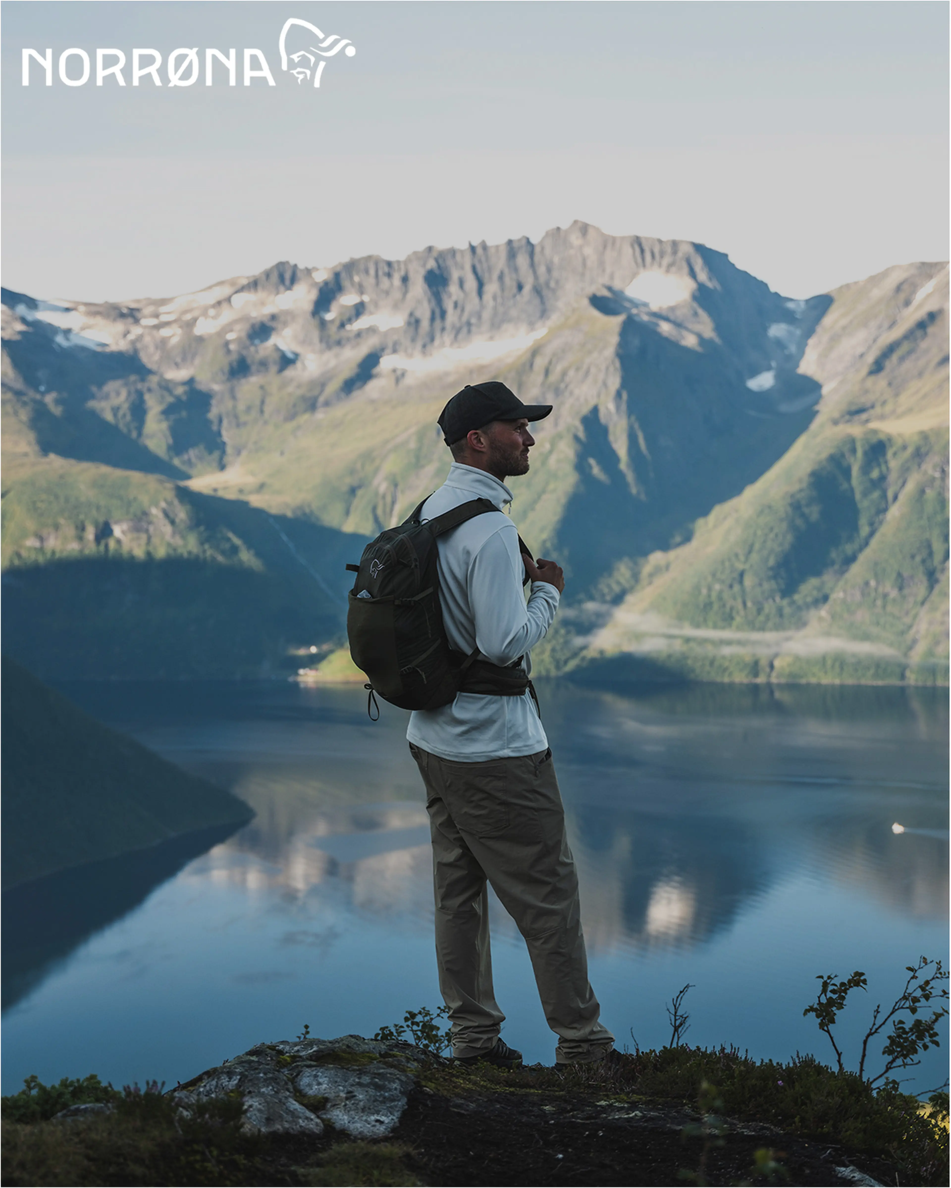A person in hiking attire stands on a mountain edge, overlooking a scenic view of a lake and rugged mountains under a clear sky. With the Norrona branding in the top left corner.