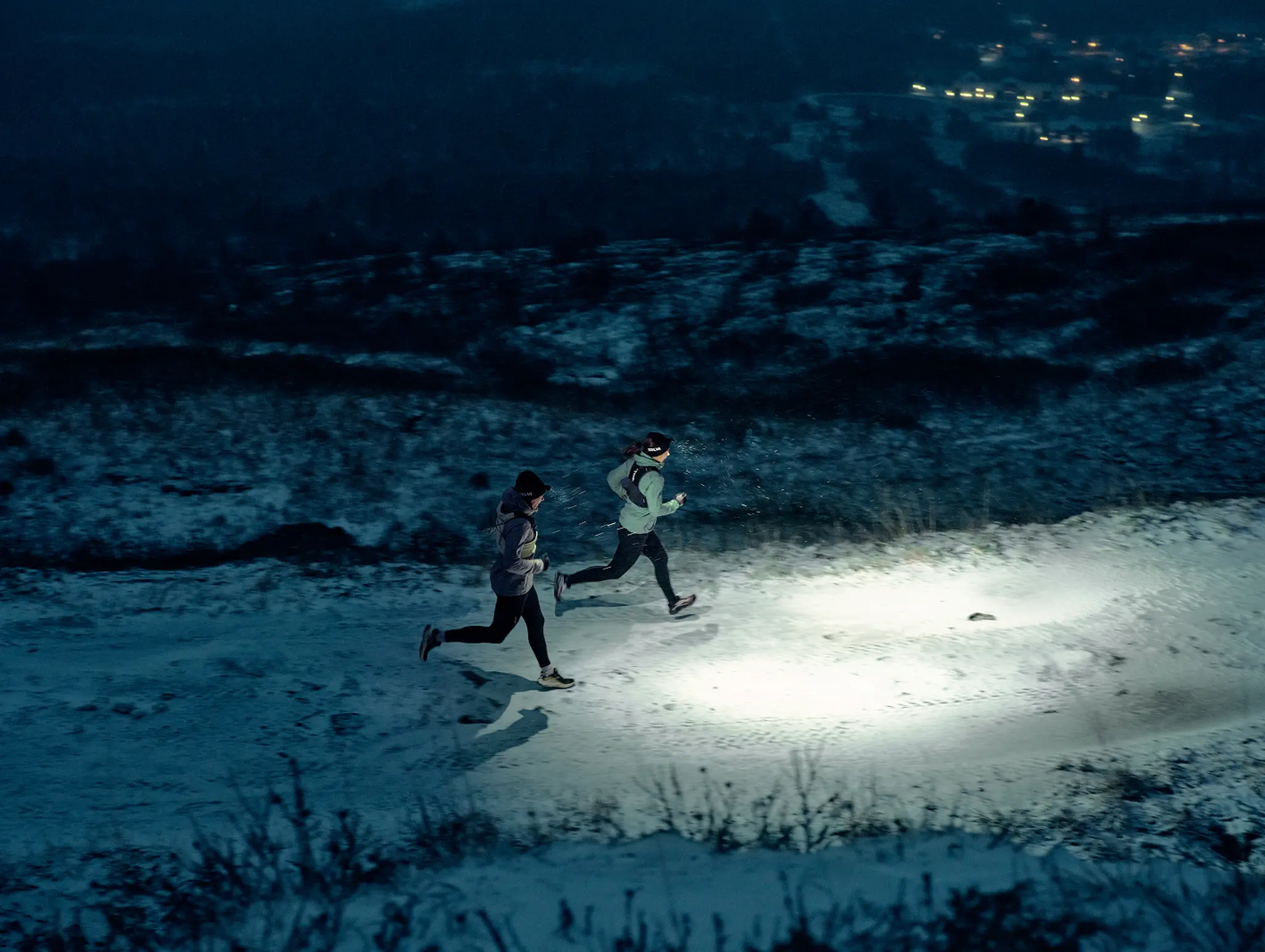 Two people running on a snowy path at night, illuminated by headlamps, with a distant view of city lights in the background.