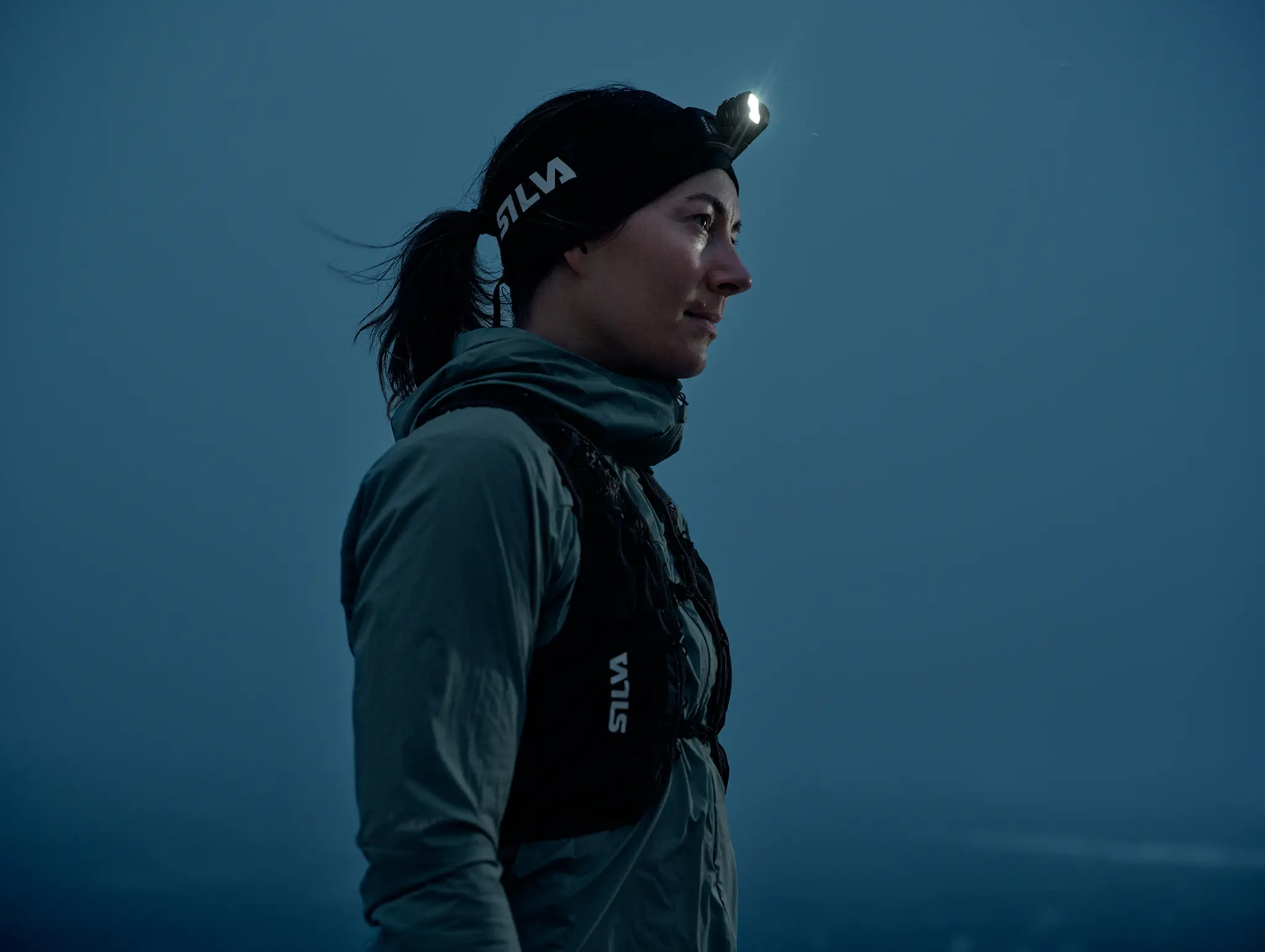 A person wearing outdoor gear and a headlamp looks into the distance at dusk, with a serene blue sky in the background.