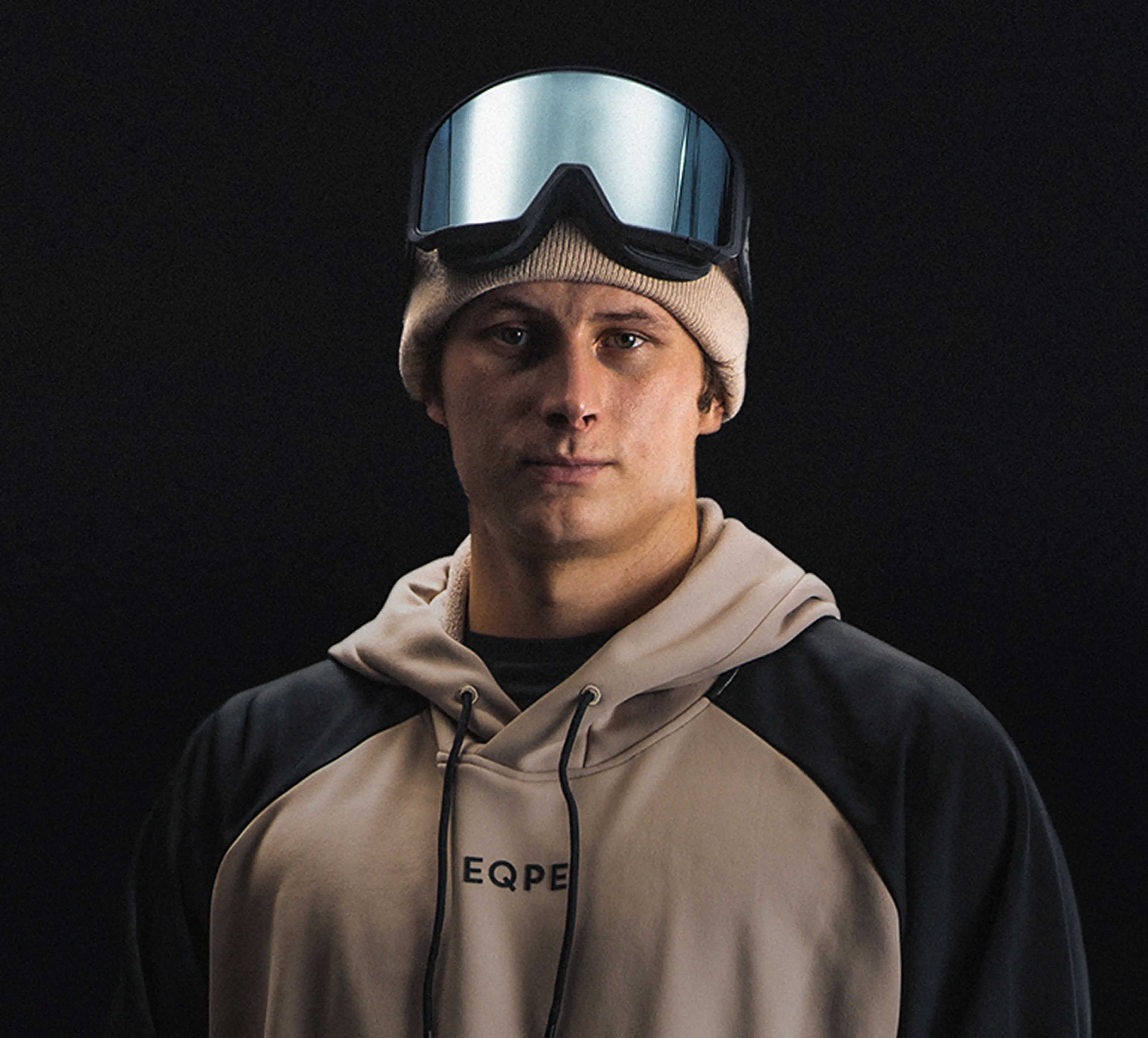 Oliwer Magnusson wearing a beige beanie, ski goggles, and a hoodie, standing against a dark background.