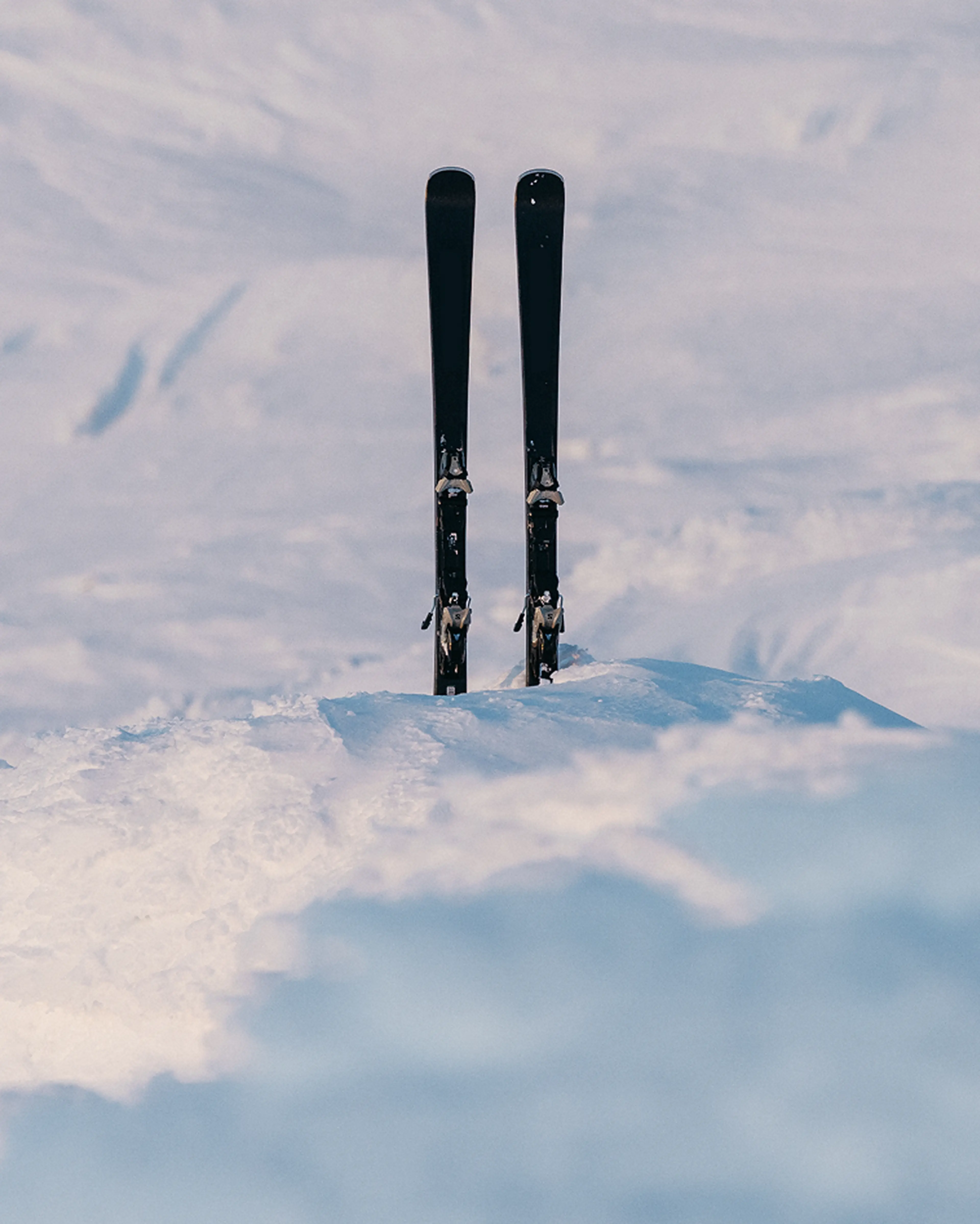 Two skis standing upright in a snow-covered landscape under a clear sky, with distant mountains in the background.