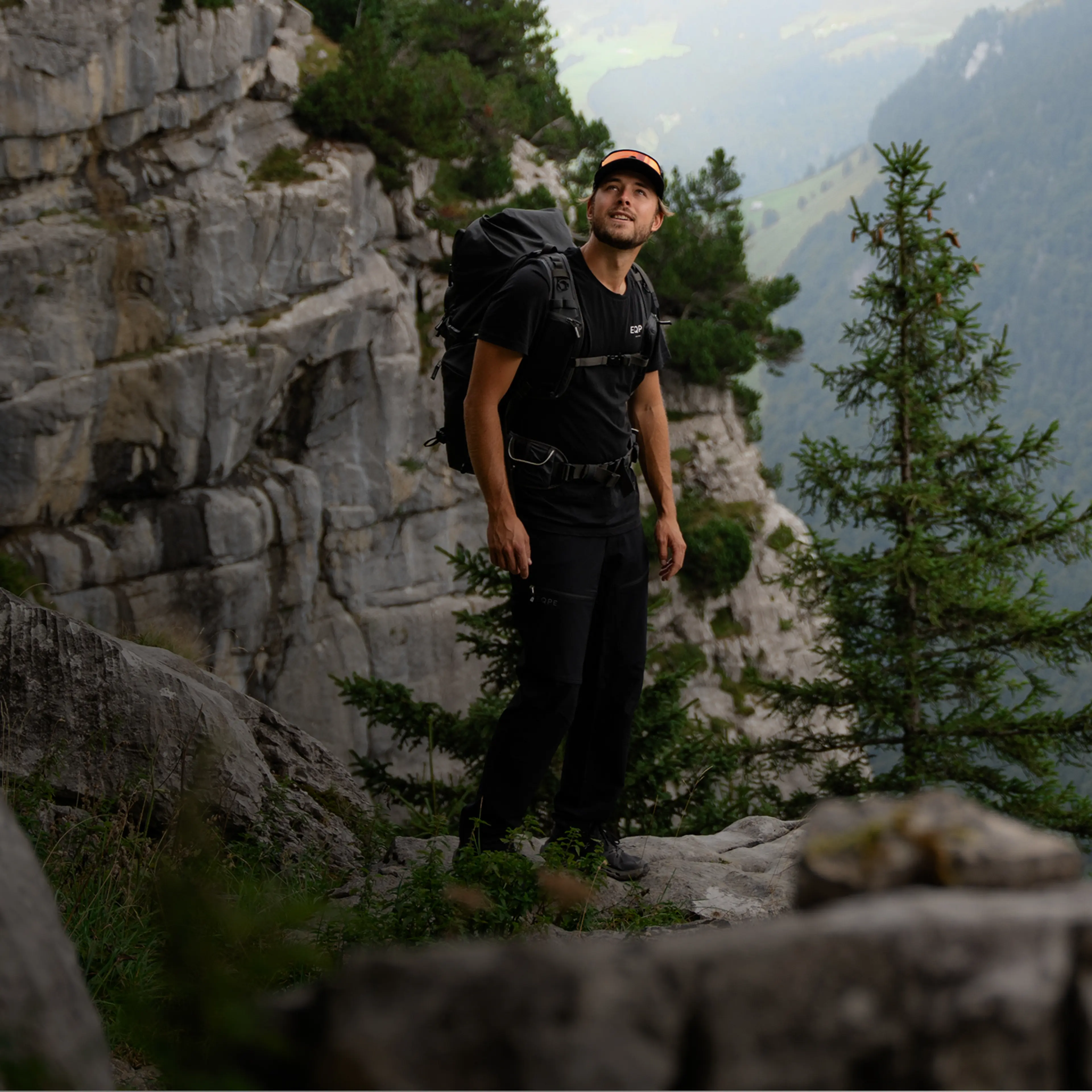 A hiker with a backpack stands on a rocky ledge, surrounded by pine trees and overlooking a mountainous landscape.