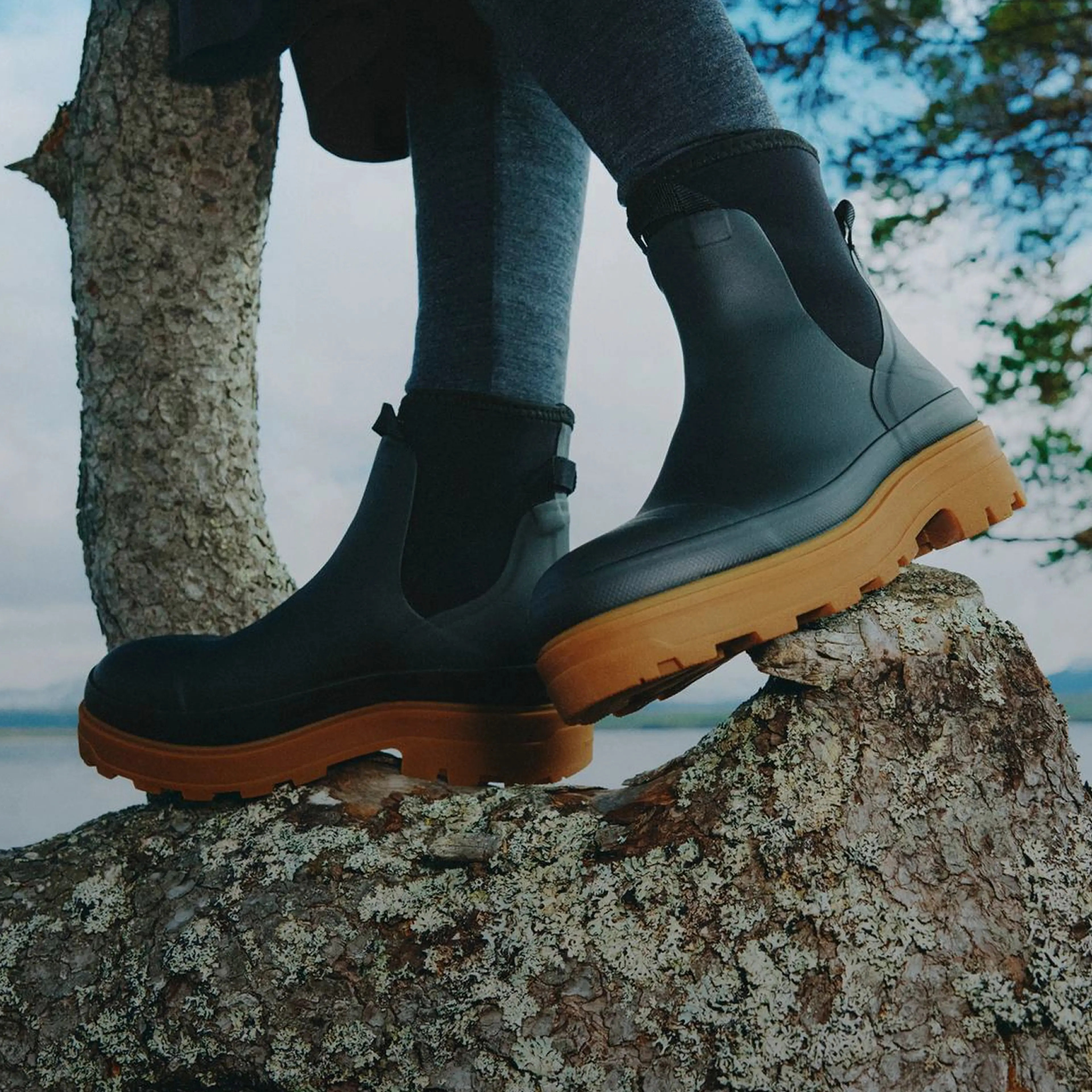 A person wearing black boots with chunky brown soles stands on a tree trunk, with a cloudy sky and water in the background.