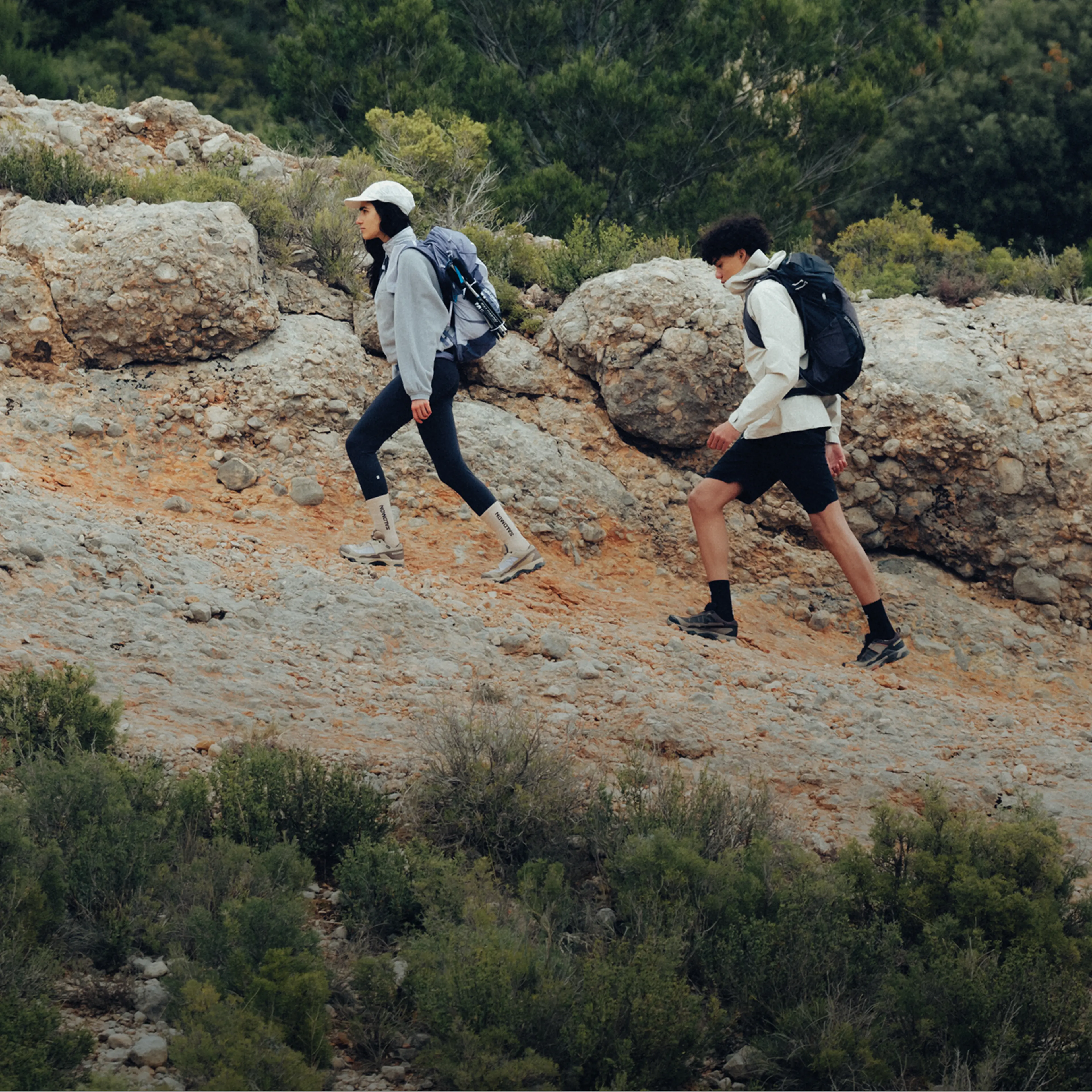Two hikers with backpacks walk up a rocky trail, surrounded by greenery, wearing casual athletic clothes and hats.