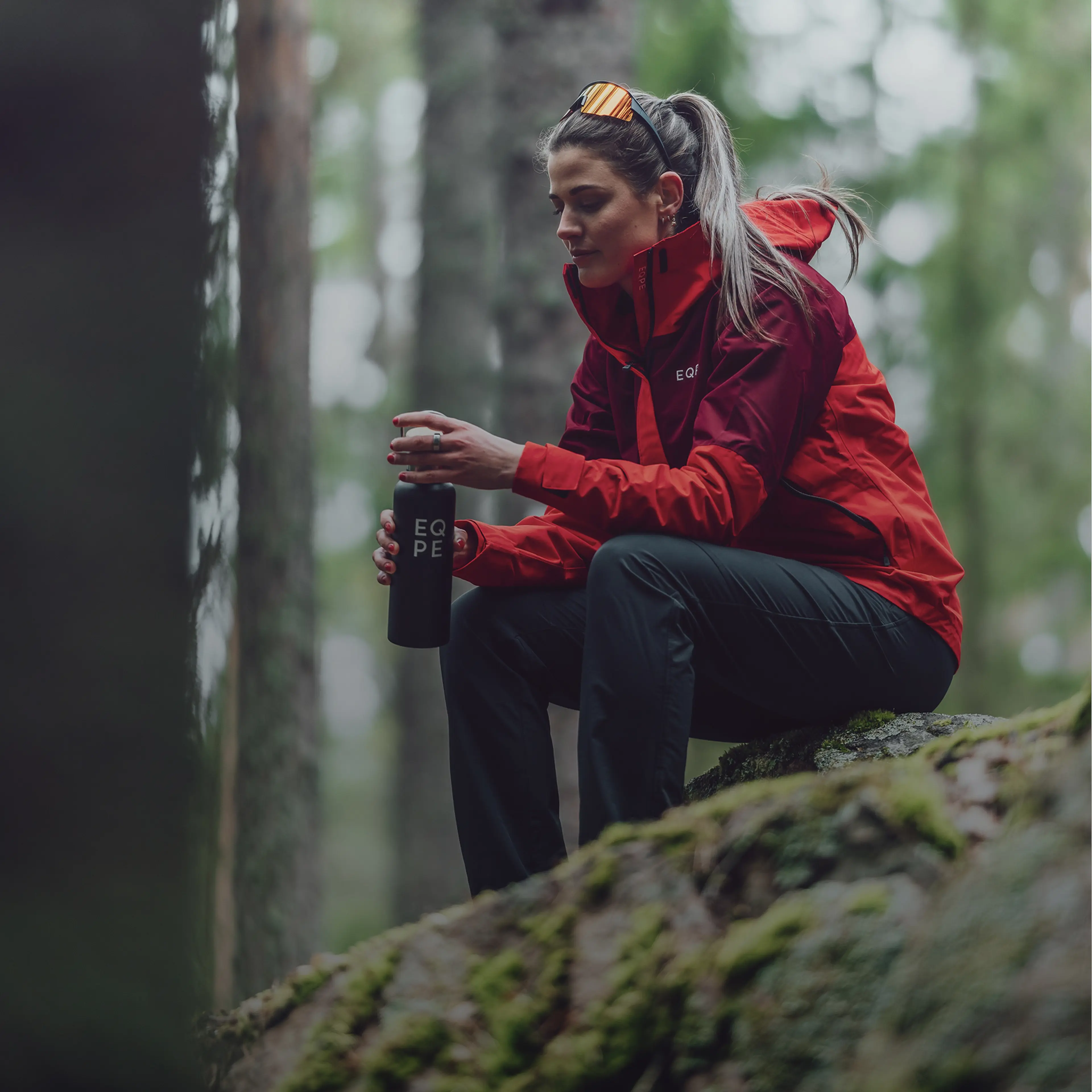 Woman wearing a red jacket, seated on a mossy rock in a forest, holding a black water bottle.