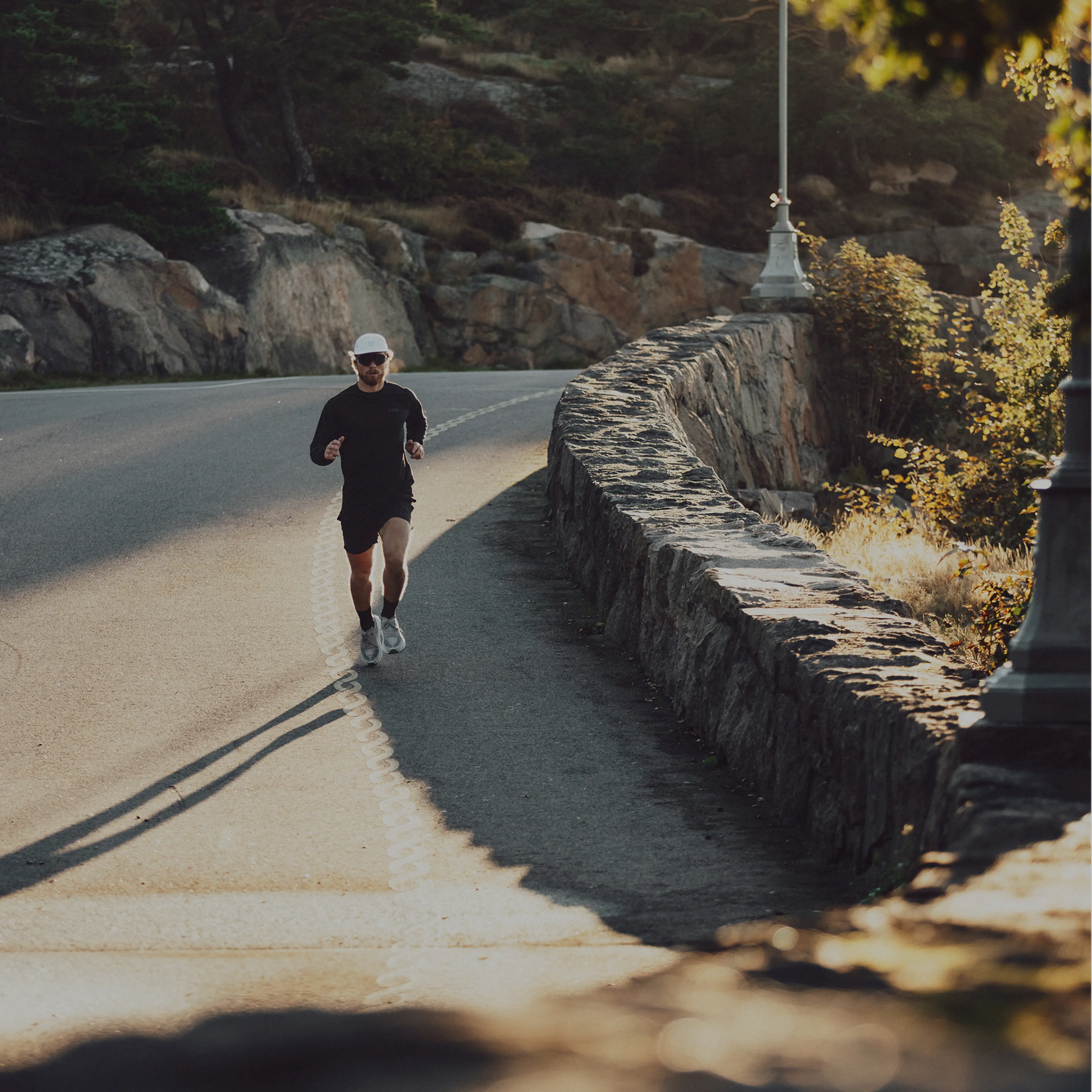 A person in sportswear and a white cap jogs along a sunny, winding road bordered by a stone wall and trees.