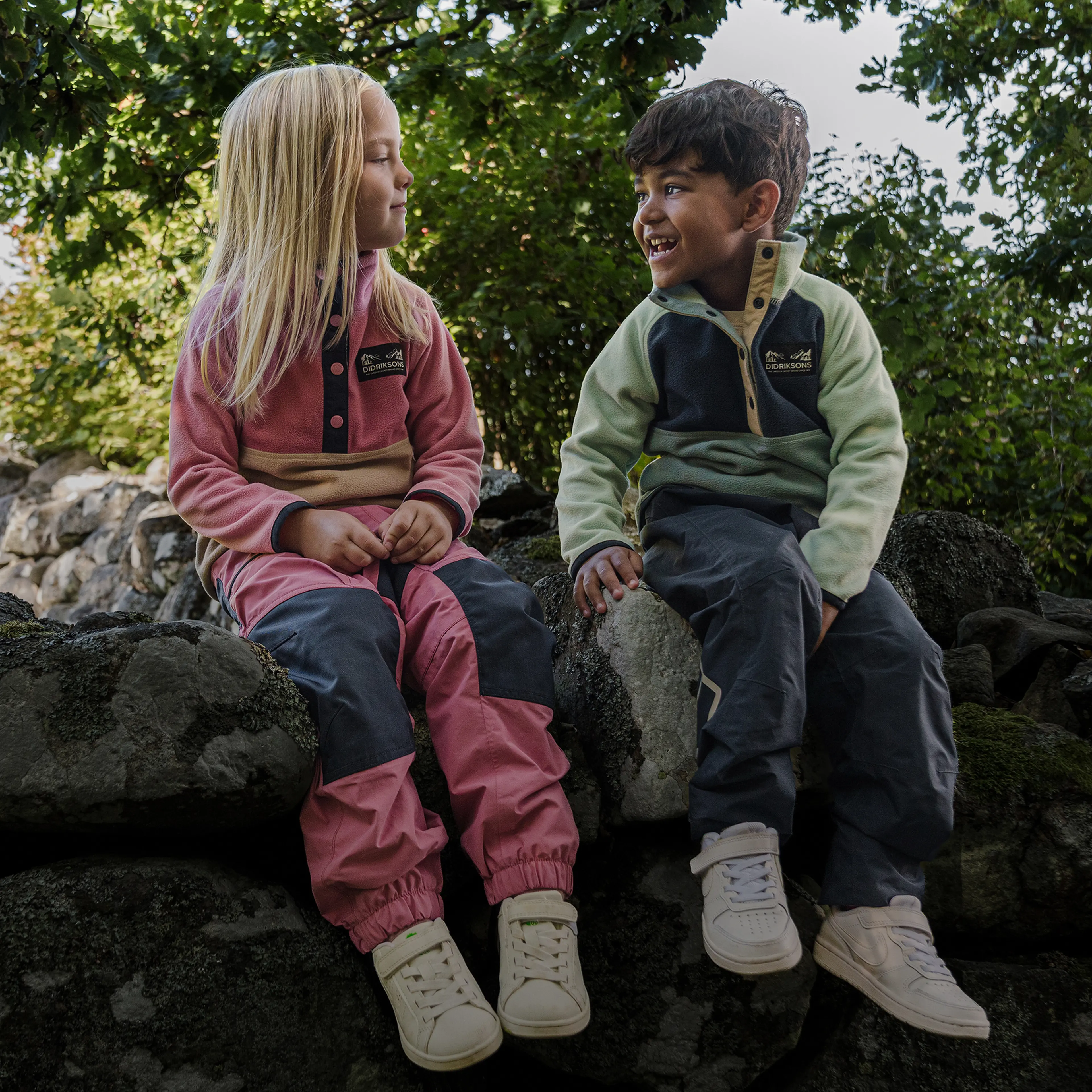 Two children sit on rocks wearing colorful outdoor clothing, surrounded by greenery, smiling and looking at each other.