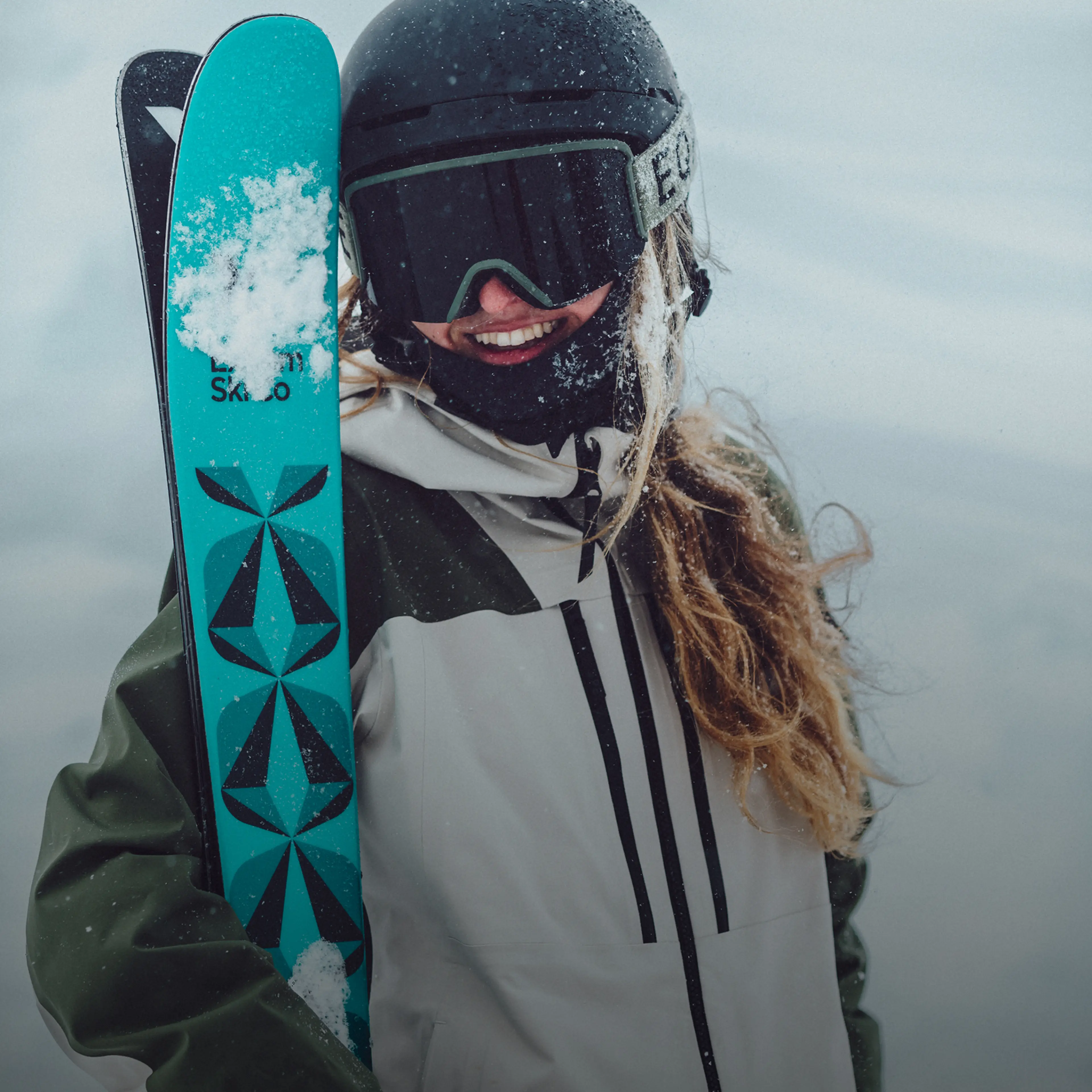 A person in ski gear smiles while holding bright turquoise skis with a snow-covered mountain backdrop.