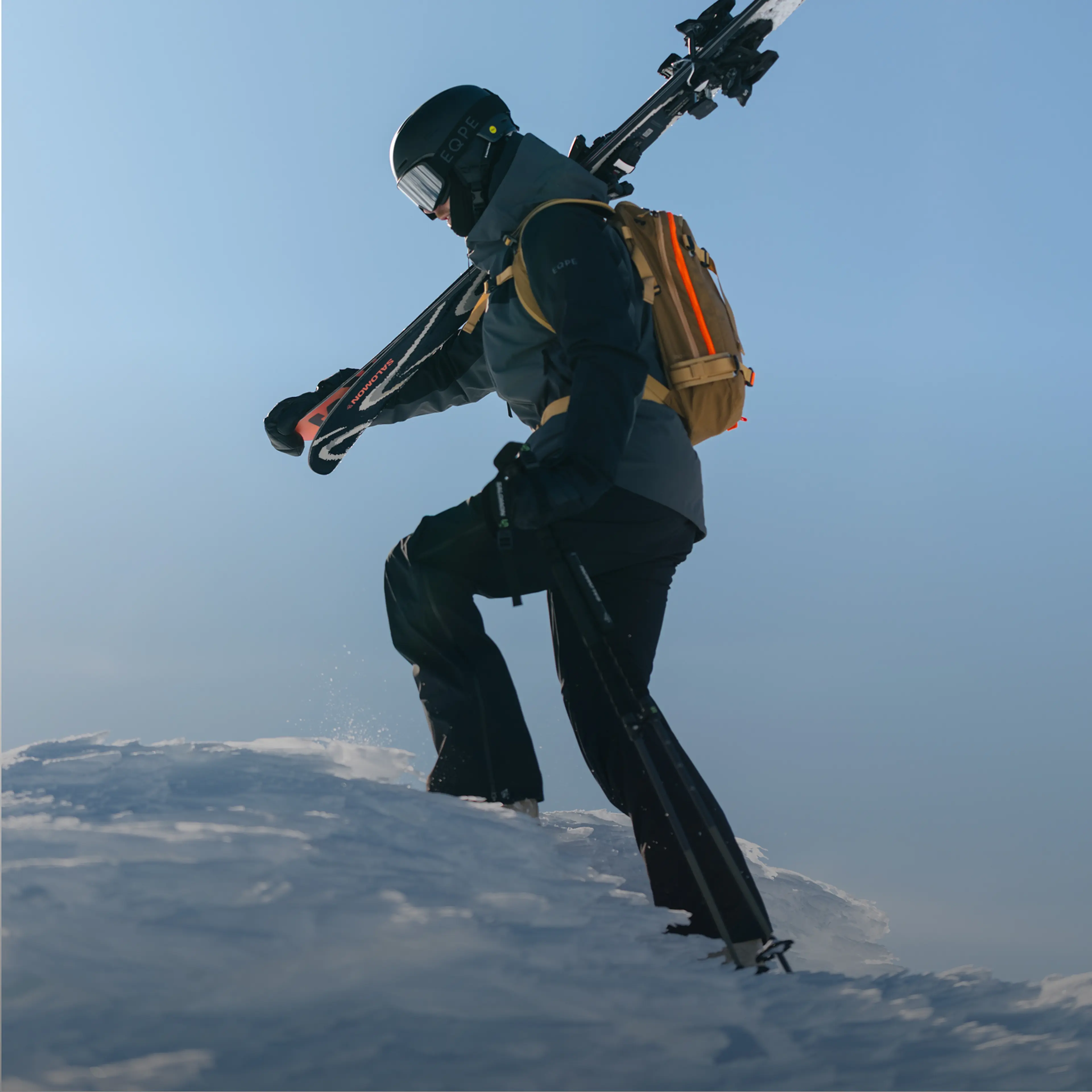 A skier in a helmet carries skis over the shoulder while walking on snow, with a bright orange backpack against a clear blue sky.