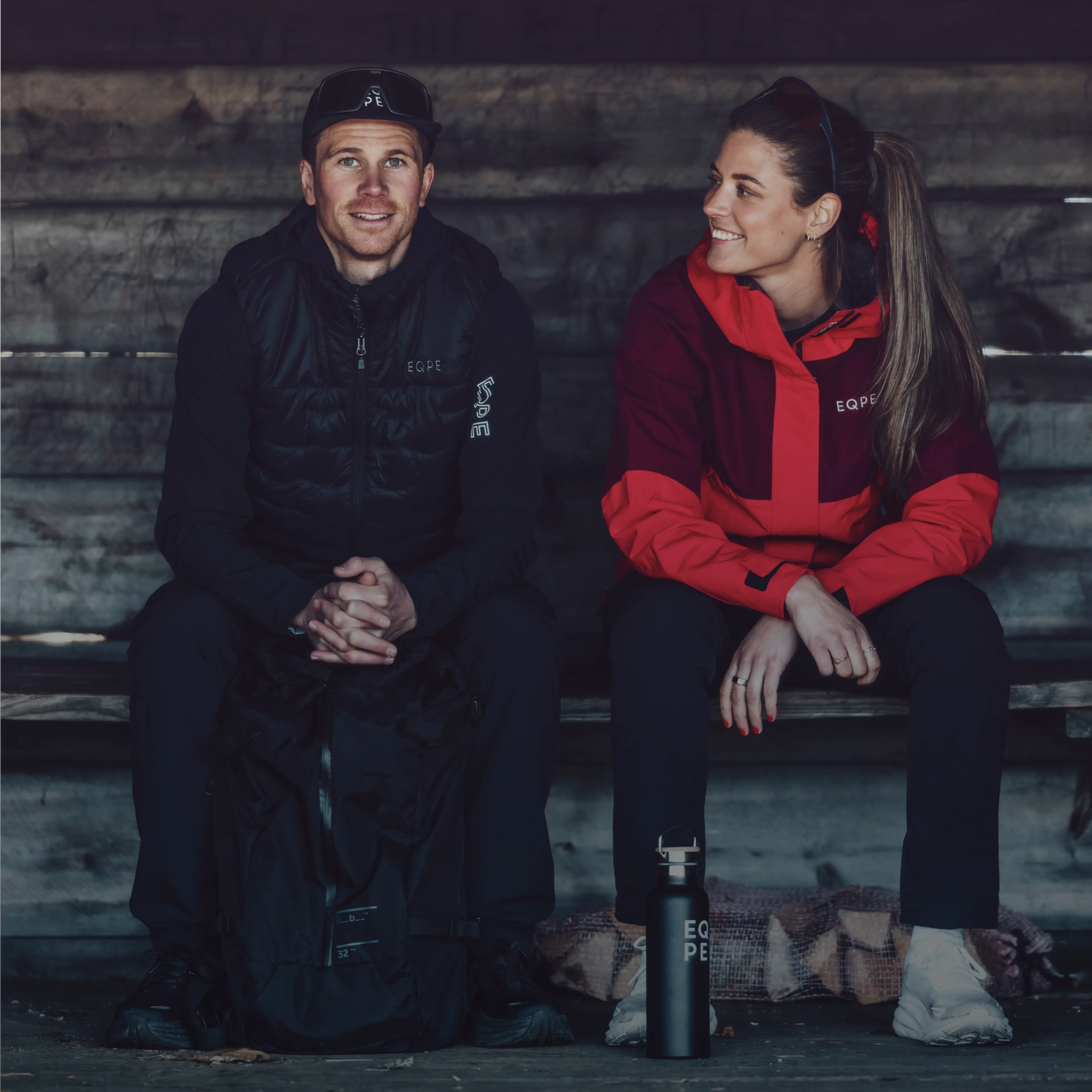Two people in outdoor gear sitting on a wooden bench, smiling at each other, with a backpack and water bottle beside them.