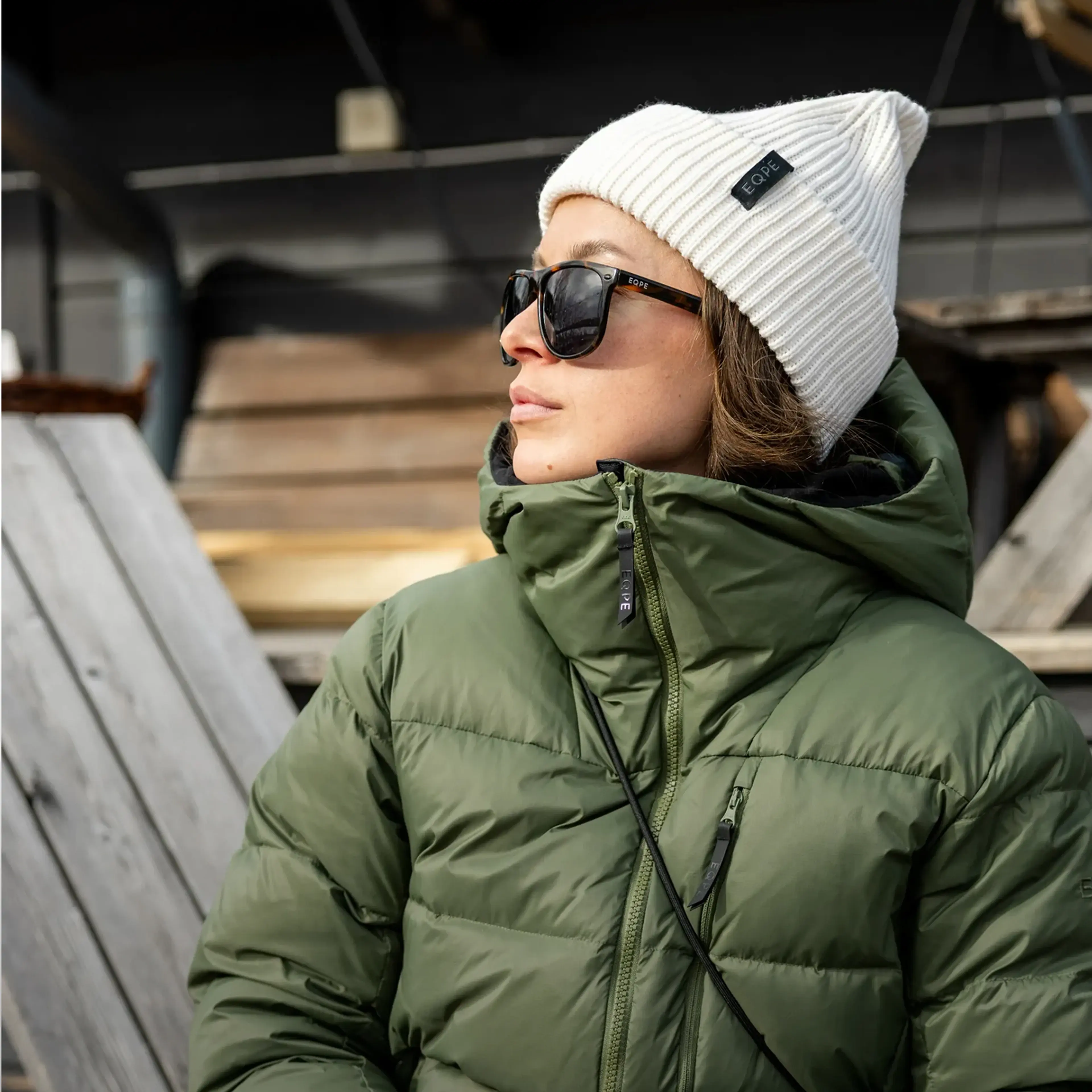 A person wearing a green puffer jacket, white beanie, and sunglasses, looking to the side outdoors.