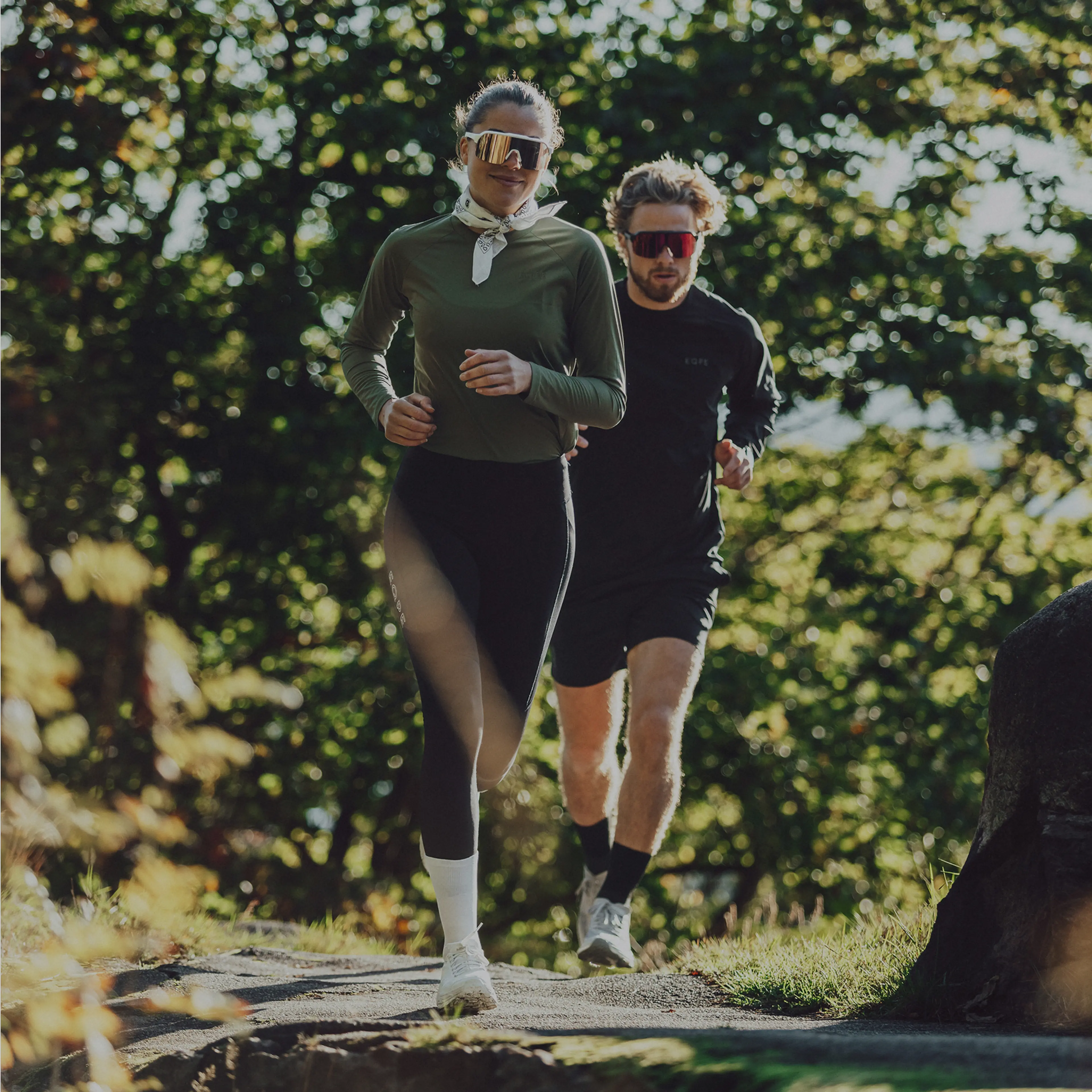 Two people jogging on a forested path, wearing athletic wear and sunglasses, with sunlight filtering through the trees.