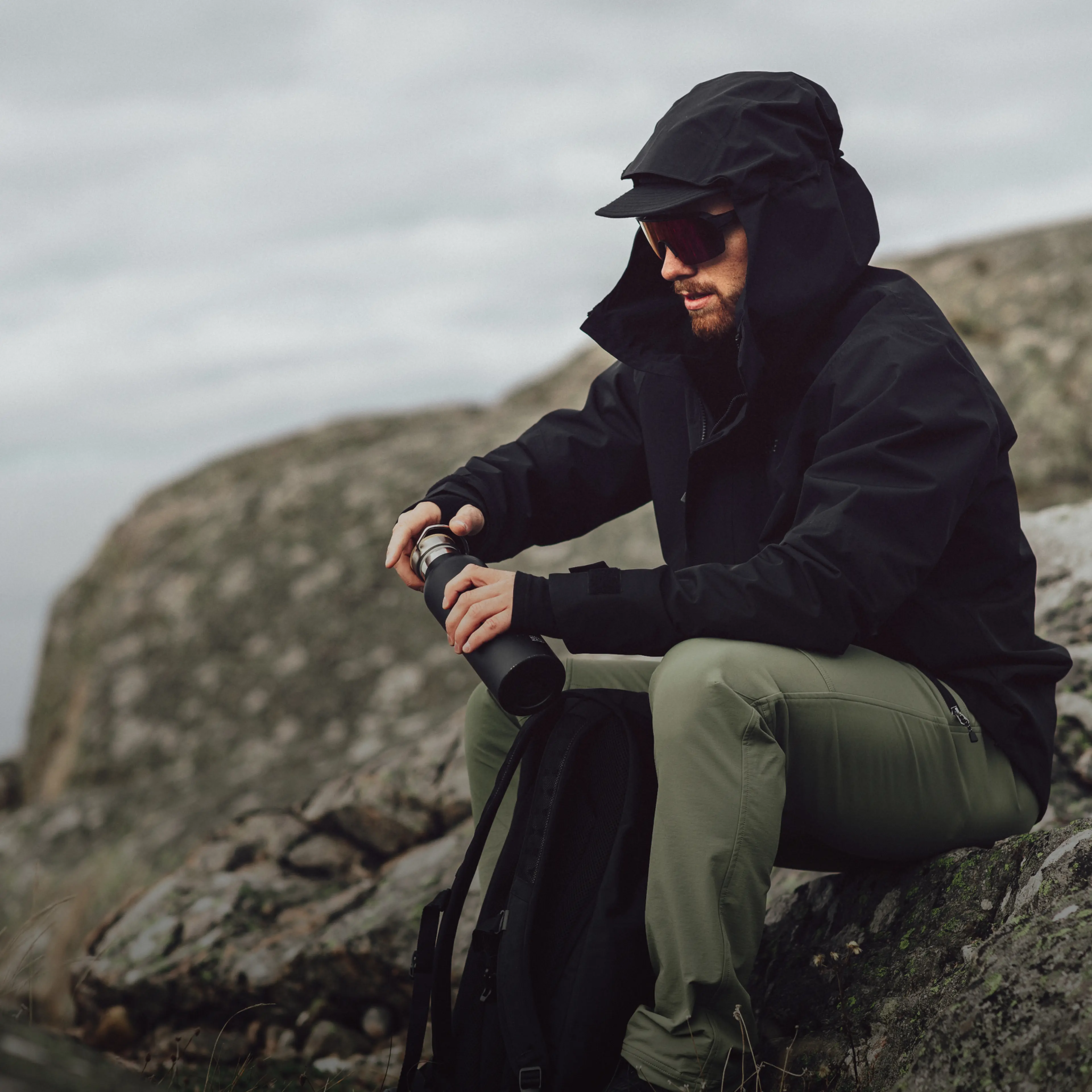 A person in outdoor gear with a hood and sunglasses sits on rocky terrain, holding a thermos and backpack, with a cloudy sky in the background.