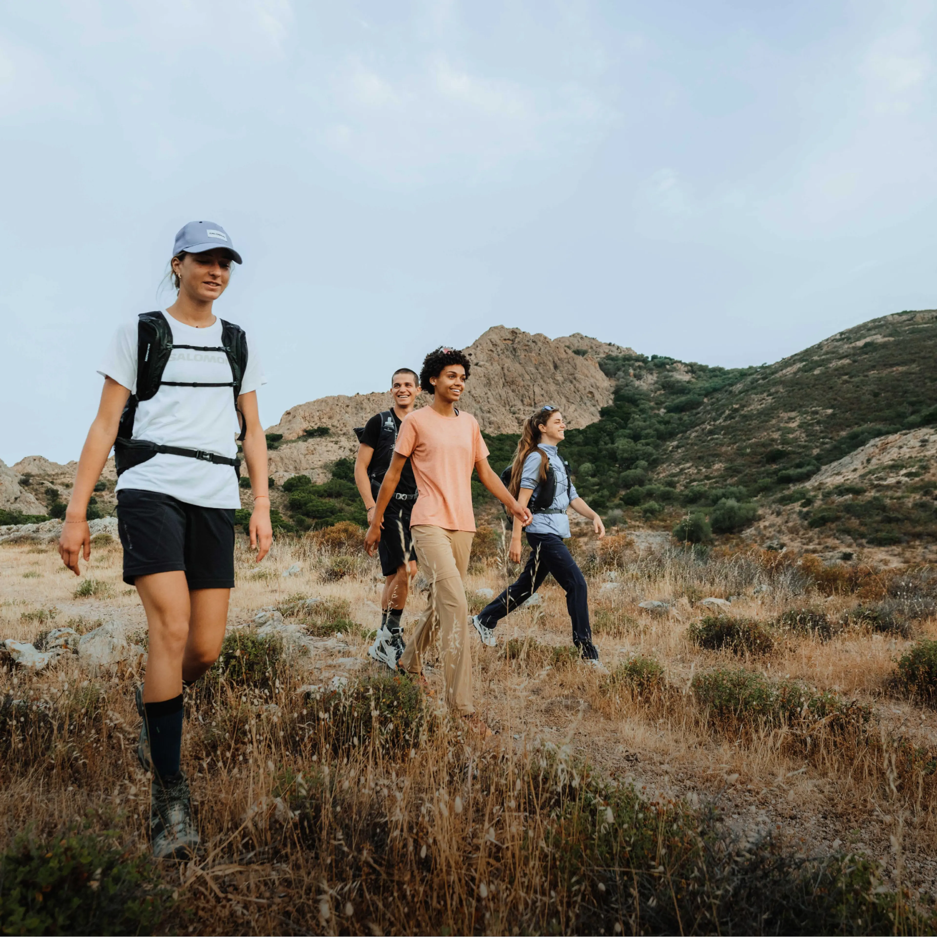 A group of four people hiking on a rocky, grassy trail in a mountainous area under a cloudy sky, wearing casual outdoor attire.