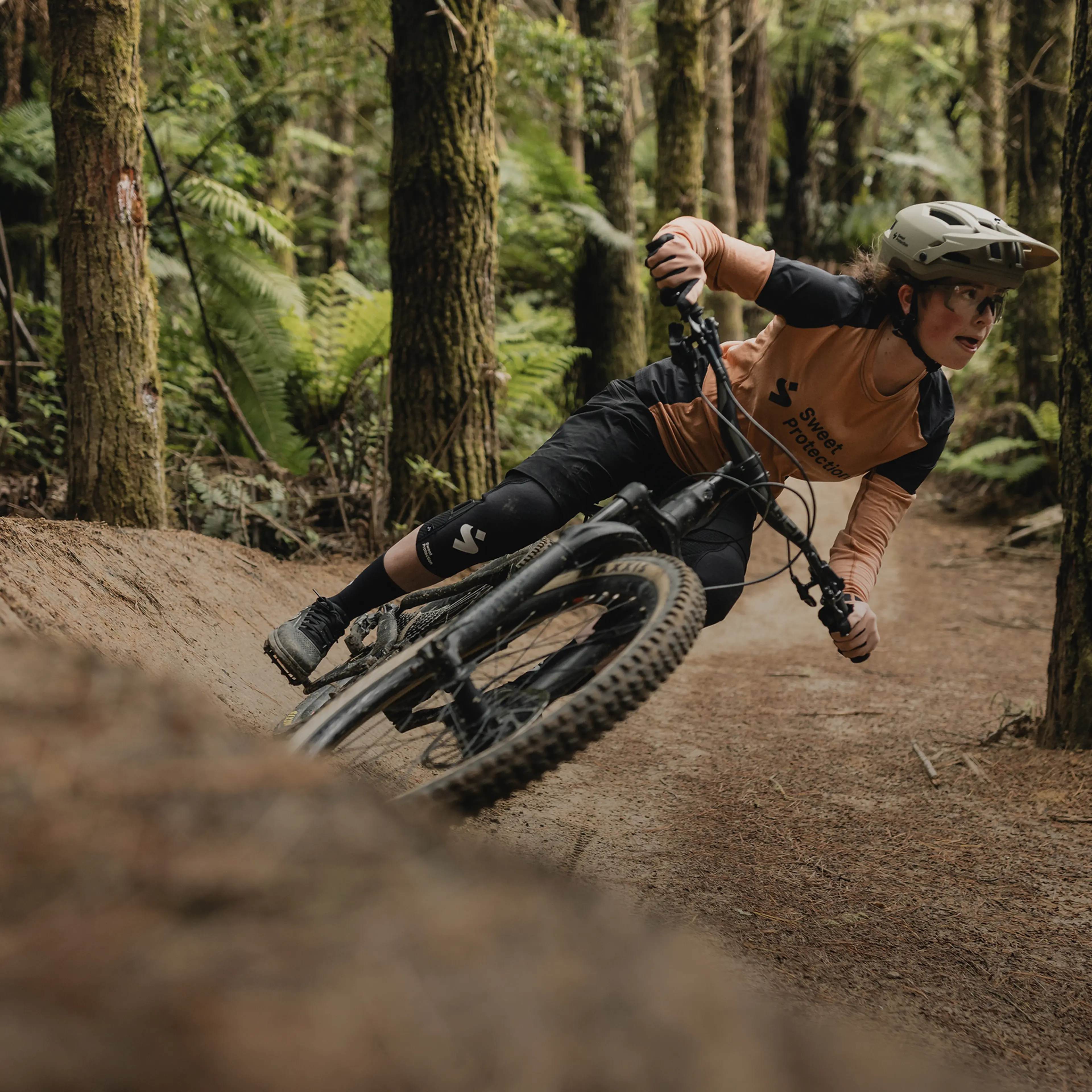 A mountain biker leans sharply while navigating a dirt trail in a dense forest, wearing protective gear and focused on the path ahead.