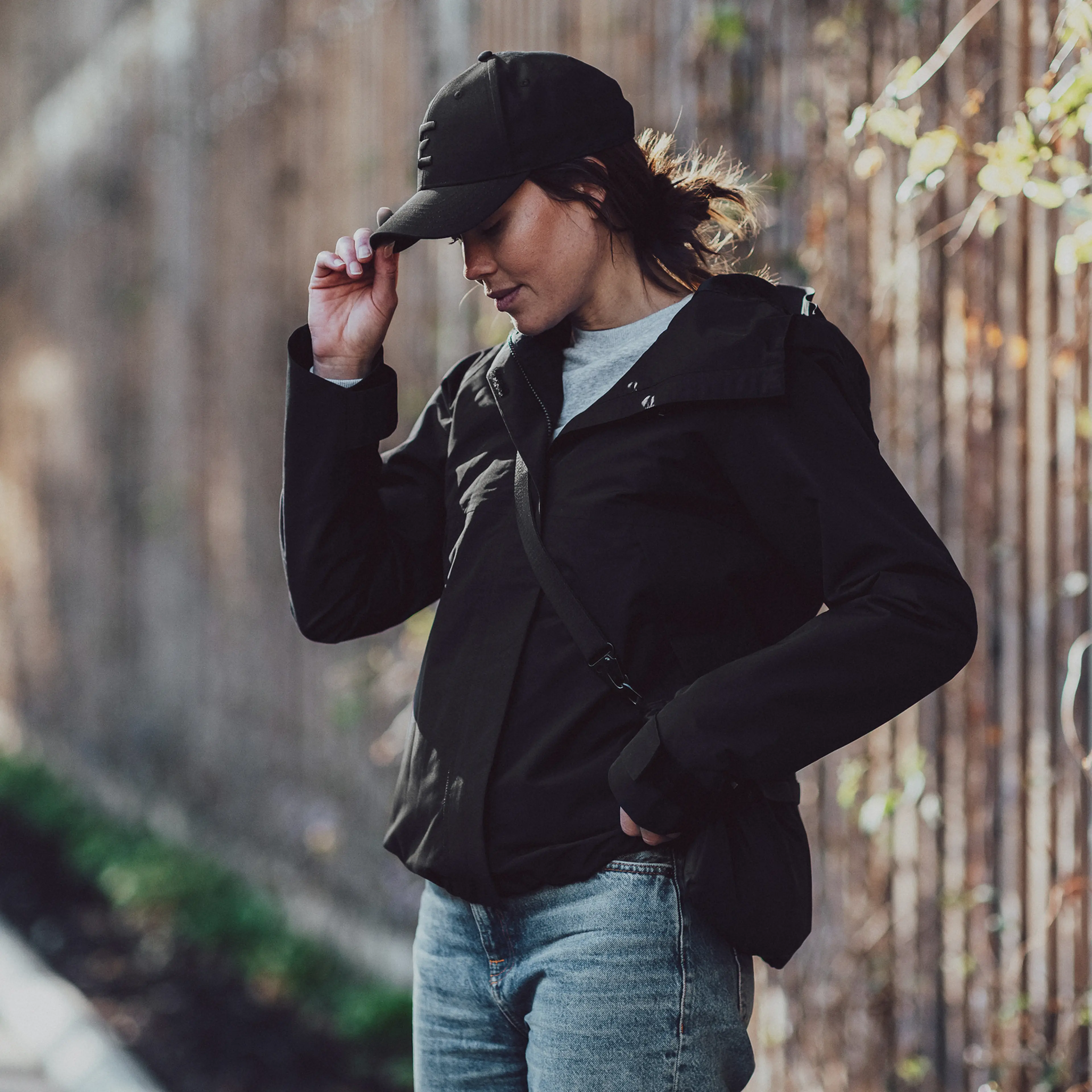 Woman in a black jacket and cap stands outdoors, adjusting her cap, with a blurred background of a wooden fence and greenery.