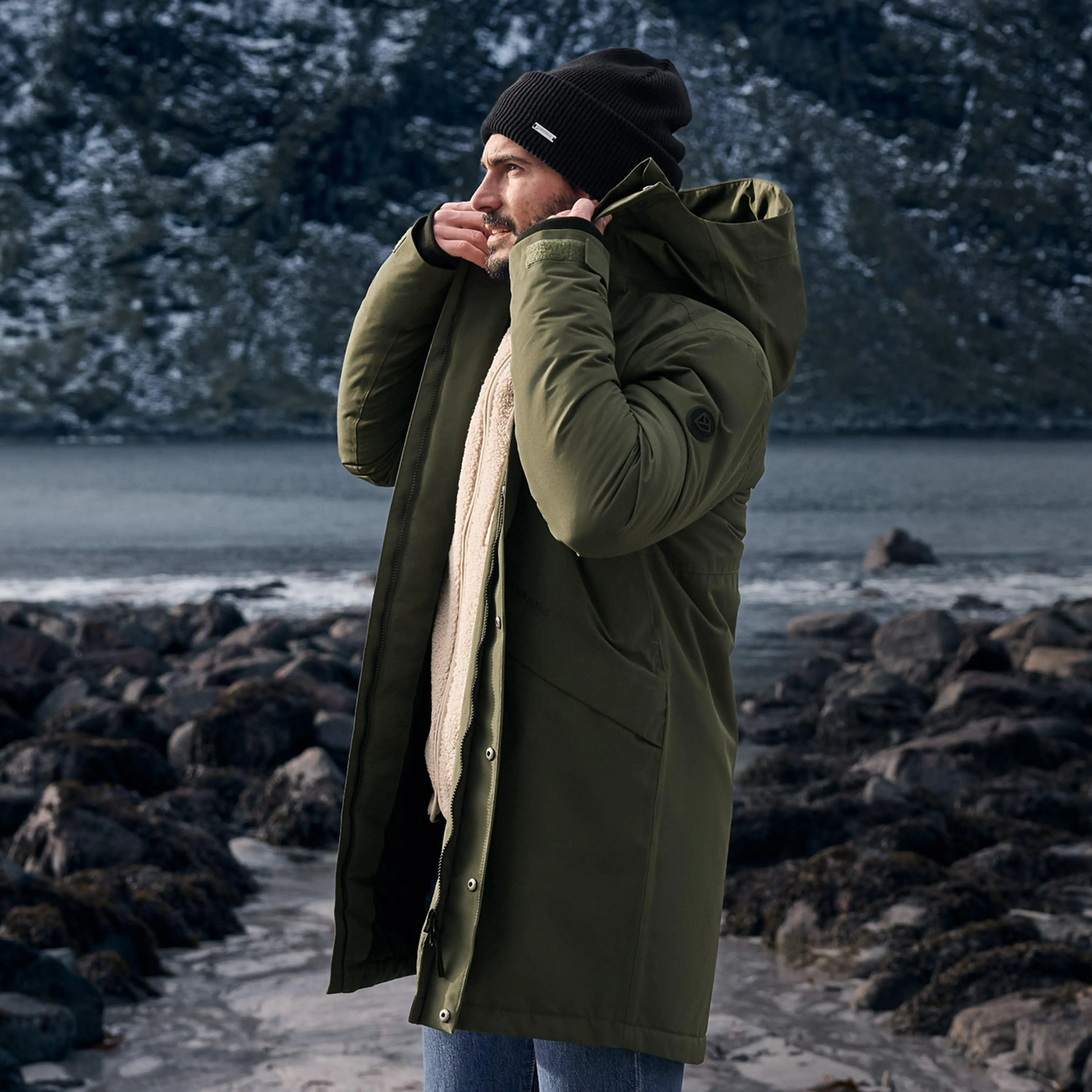 A person in a green winter coat and black beanie stands on a rocky shoreline, with a snowy mountain in the background.