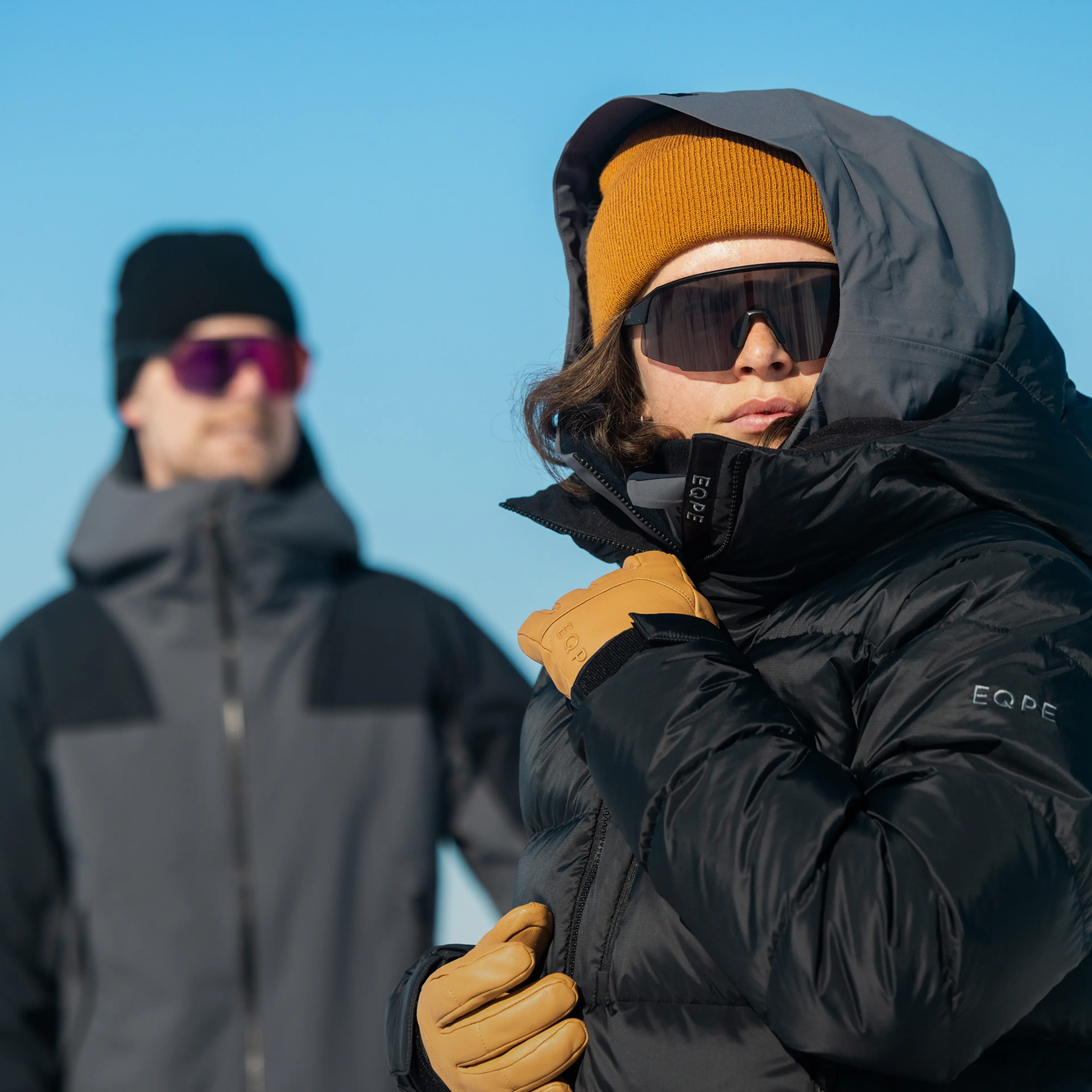 Two individuals in winter attire and sunglasses, one wearing an orange beanie and black puffer jacket, stand against a clear blue sky.