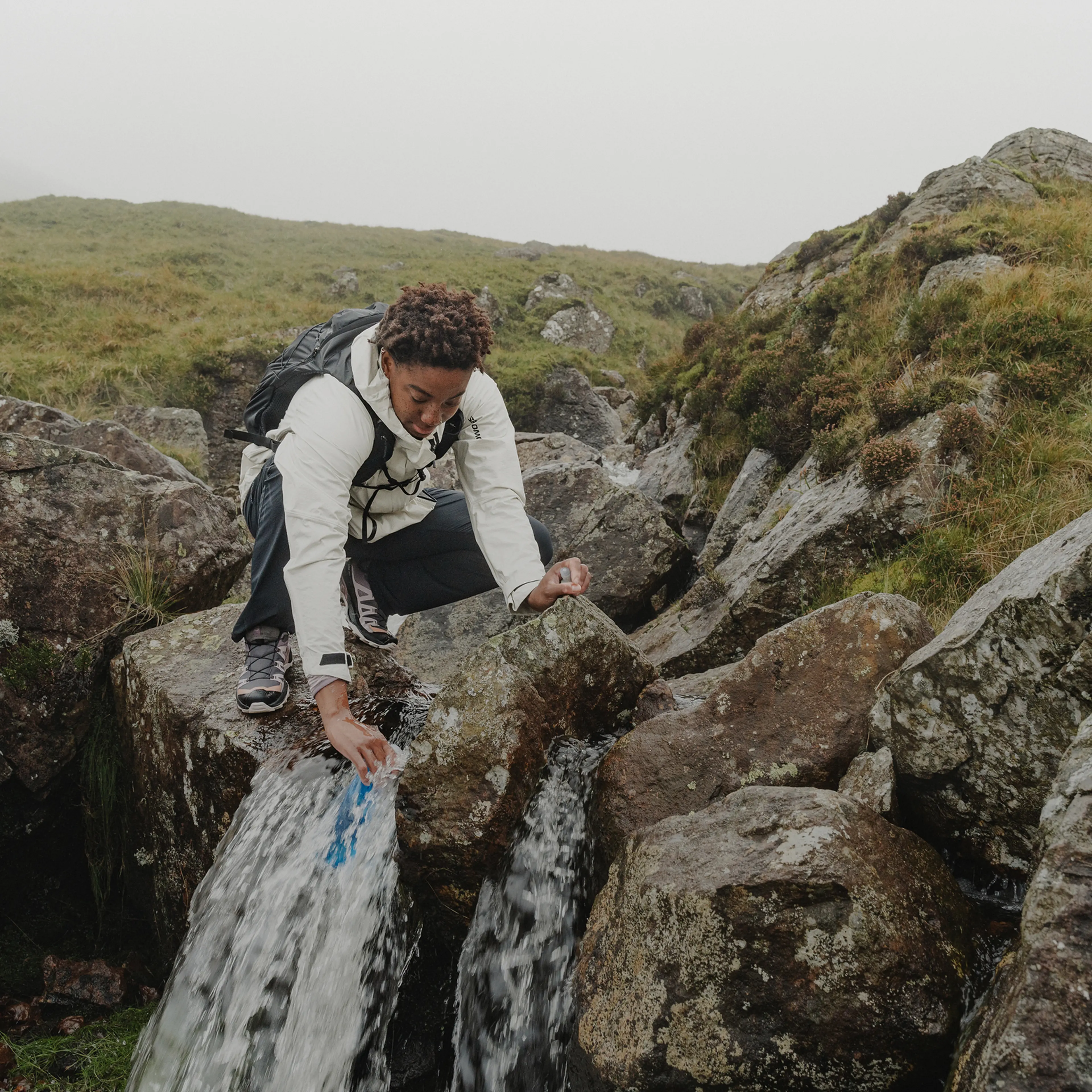 A person in a white jacket and backpack collects water from a stream in a grassy, rocky landscape.