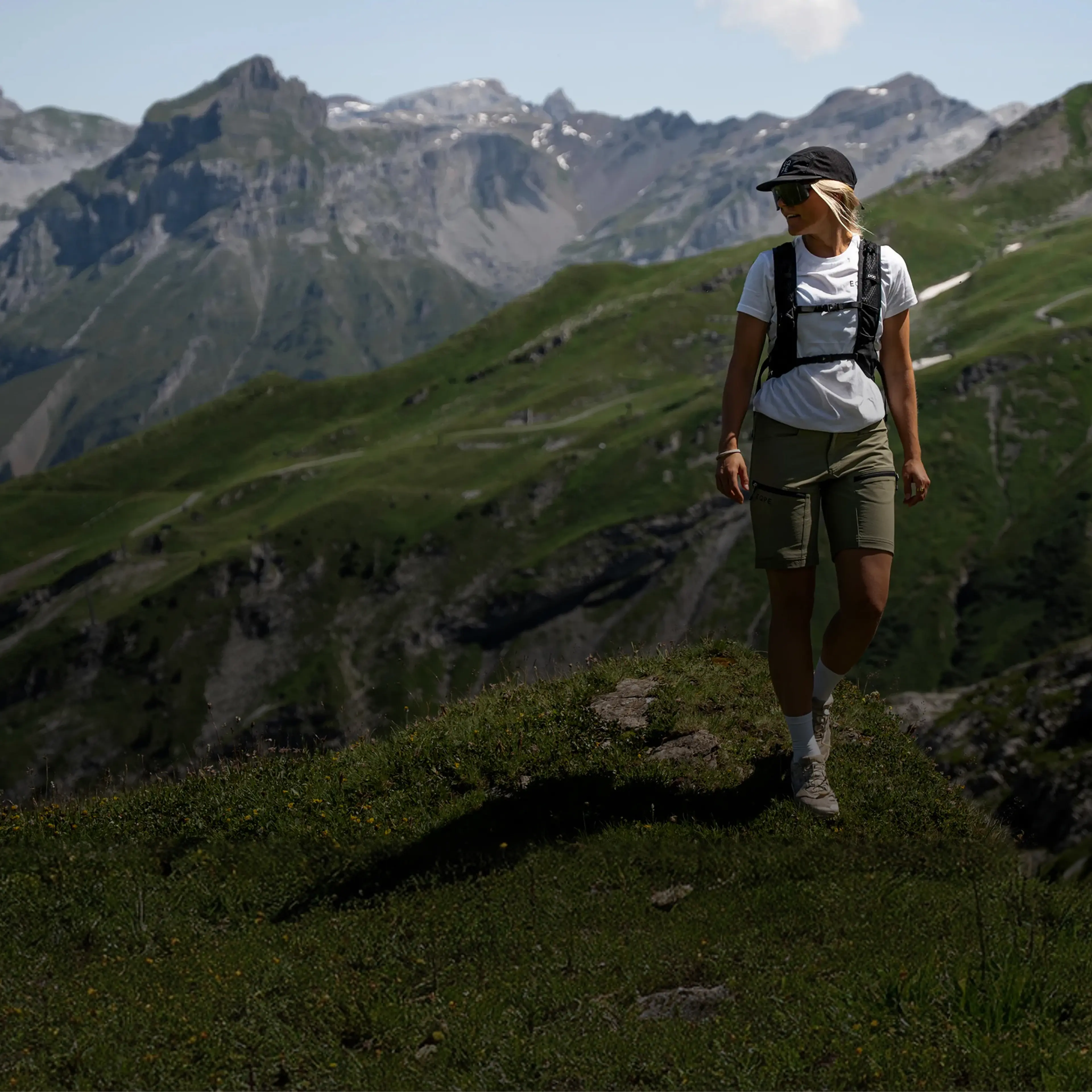 A person hikes along a grassy mountain ridge wearing a cap and backpack, with rocky peaks and lush valleys in the background.
