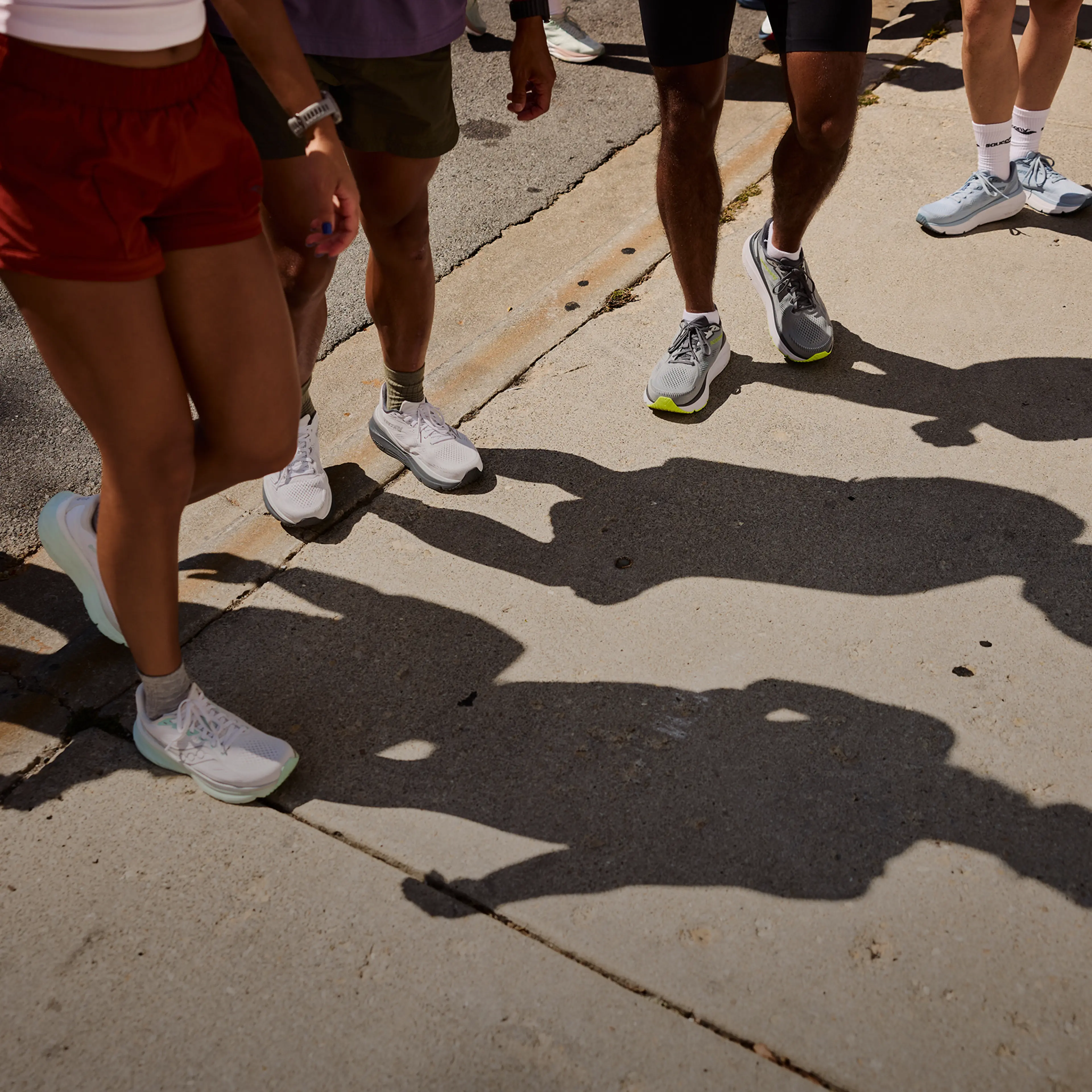 A group of people walking on a sunlit street, with their shadows cast prominently on the sidewalk. They wear casual athletic shoes.