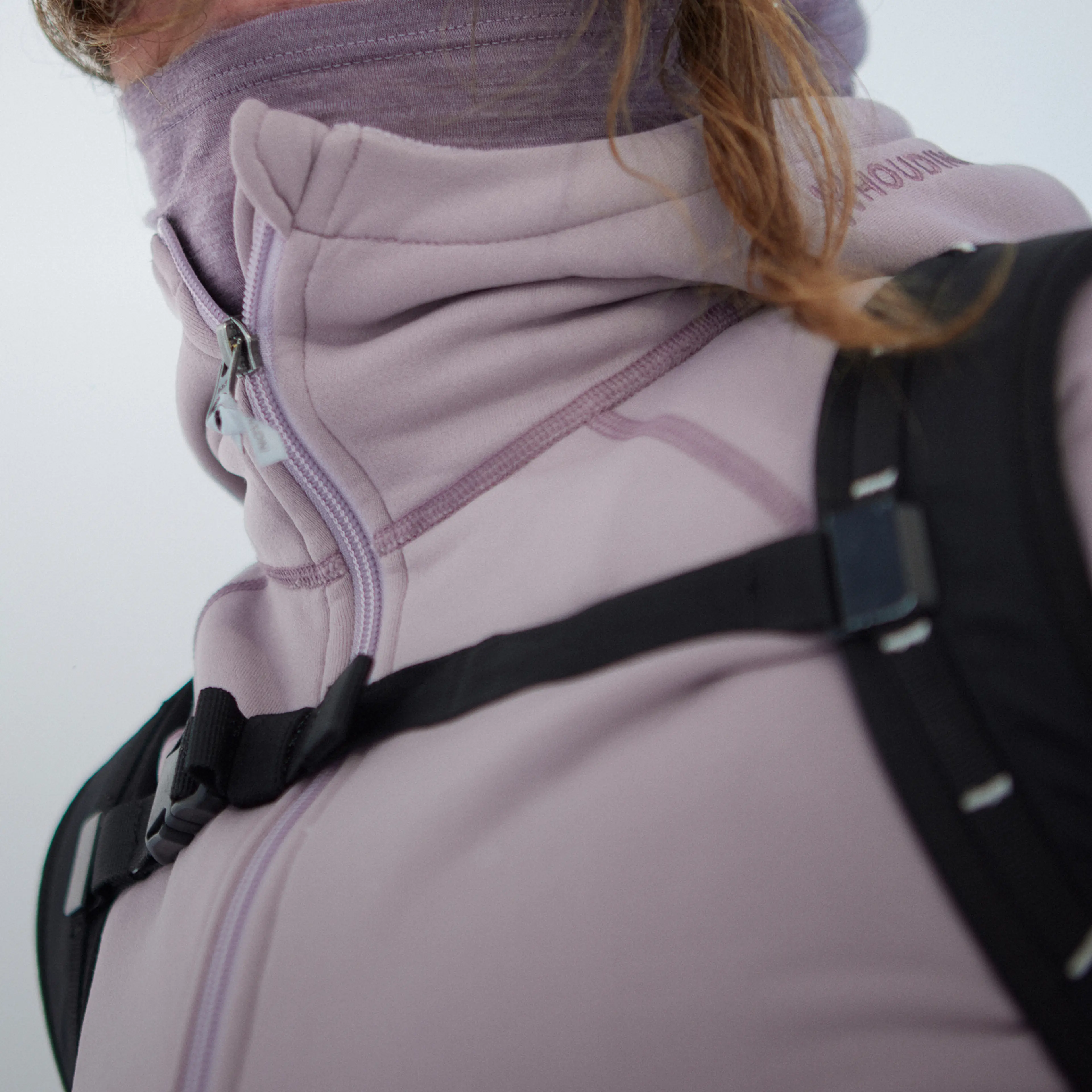 Person wearing a light purple jacket with a high collar and a backpack strap across the chest, partially covering their face.