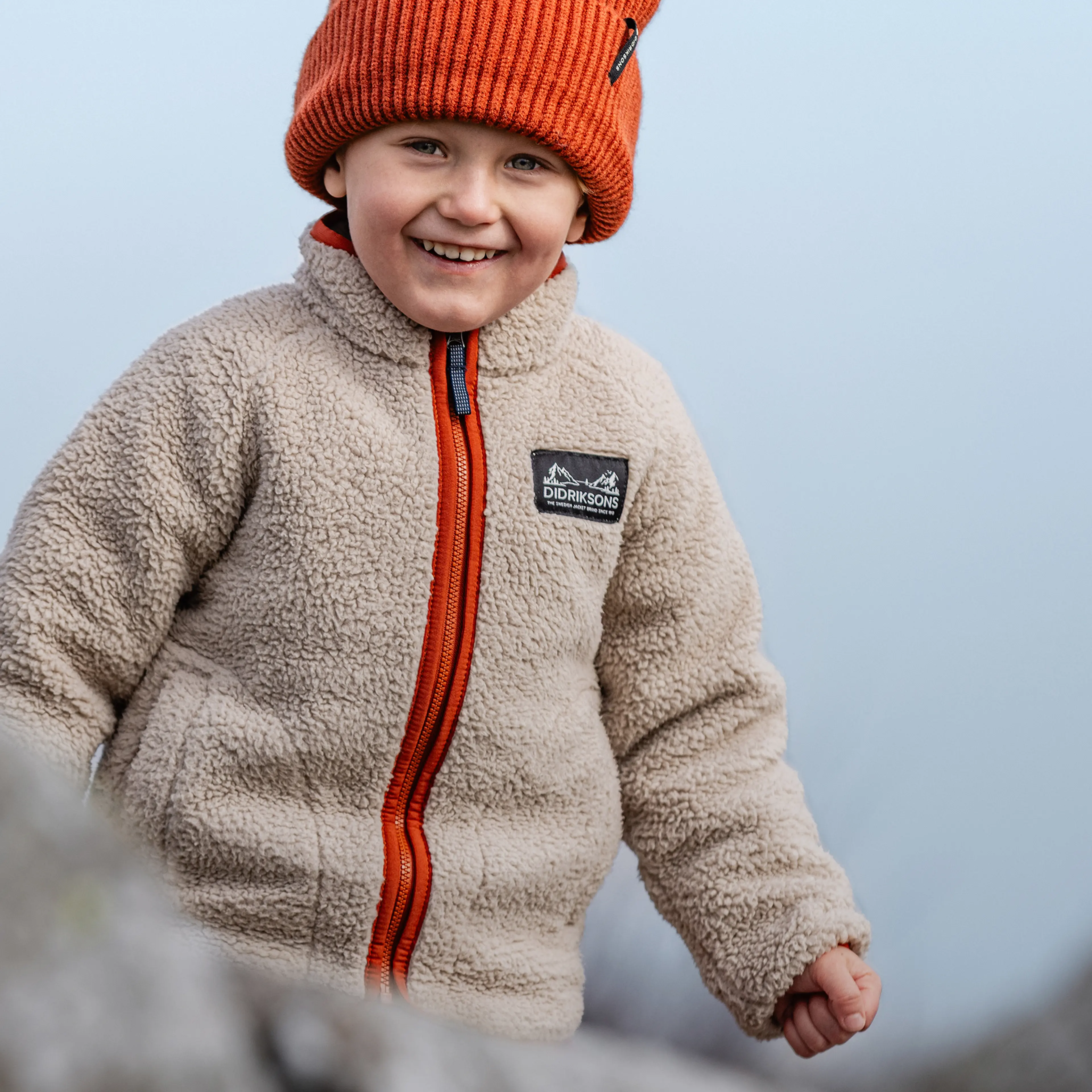 A smiling child in a beige fleece jacket and orange knit hat stands outdoors against a light blue background.
