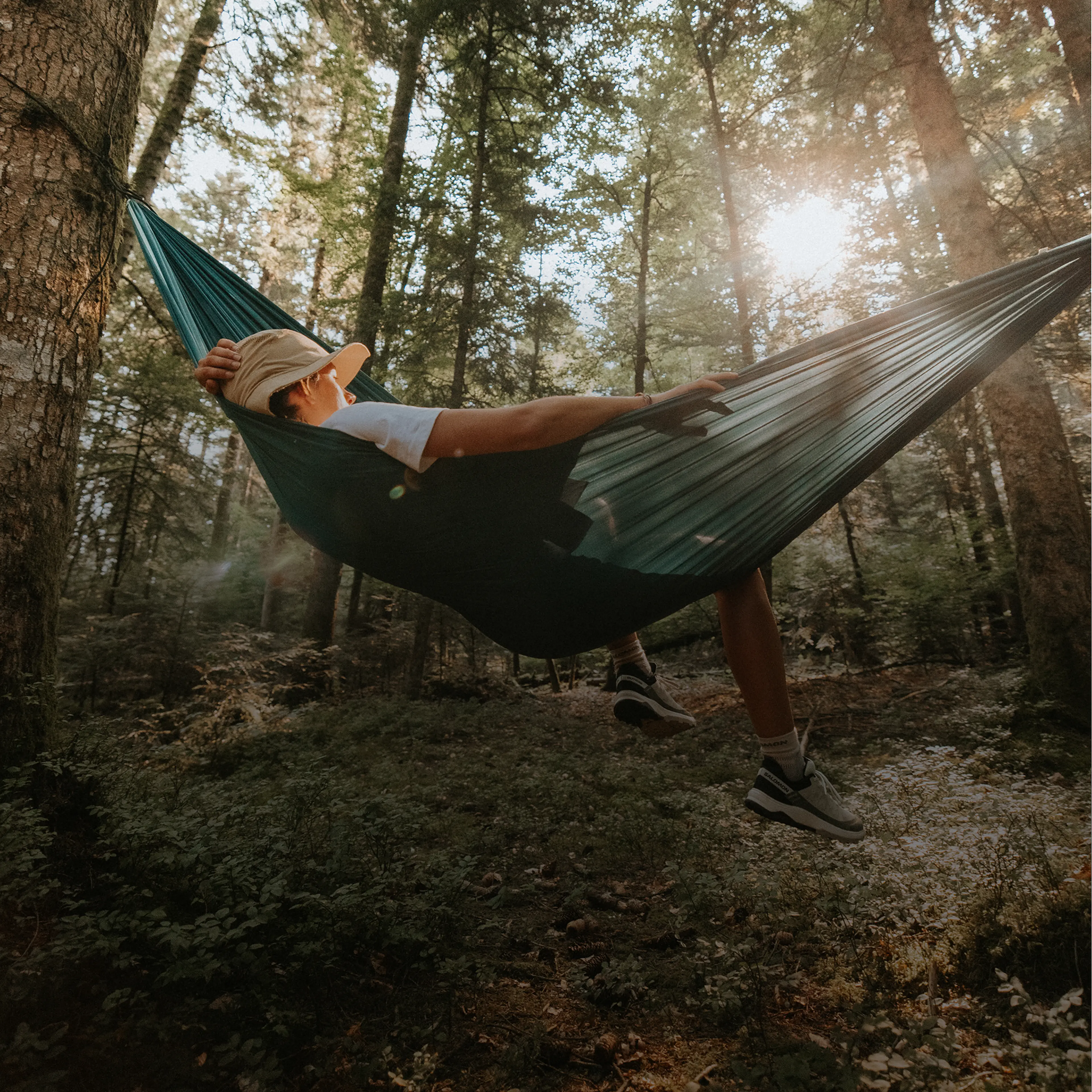 Person relaxing in a green hammock in a sunlit forest, wearing a hat and sneakers, surrounded by tall trees and greenery.