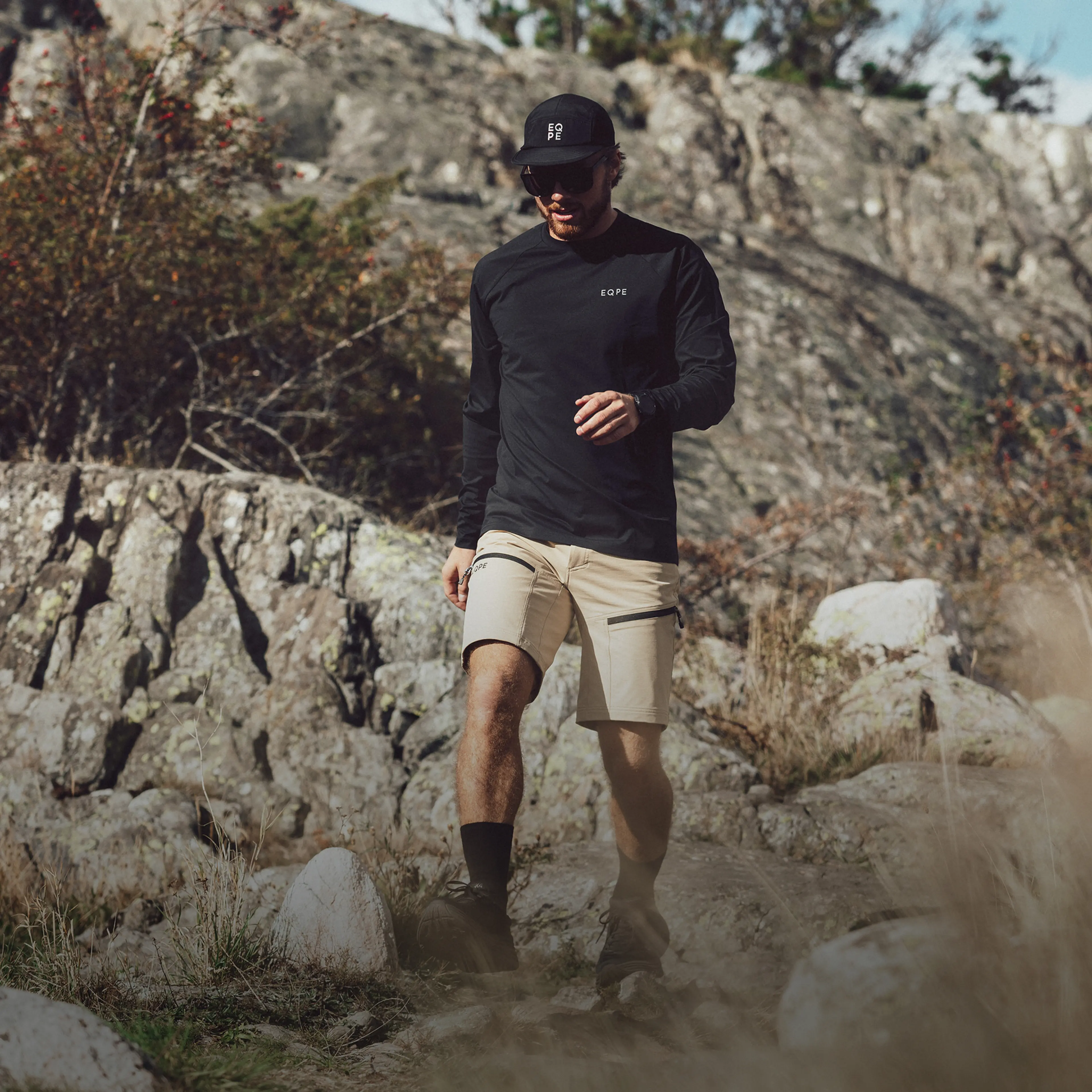 Man in outdoor clothing hiking on a rocky trail, wearing a black cap and sunglasses, surrounded by rugged terrain.
