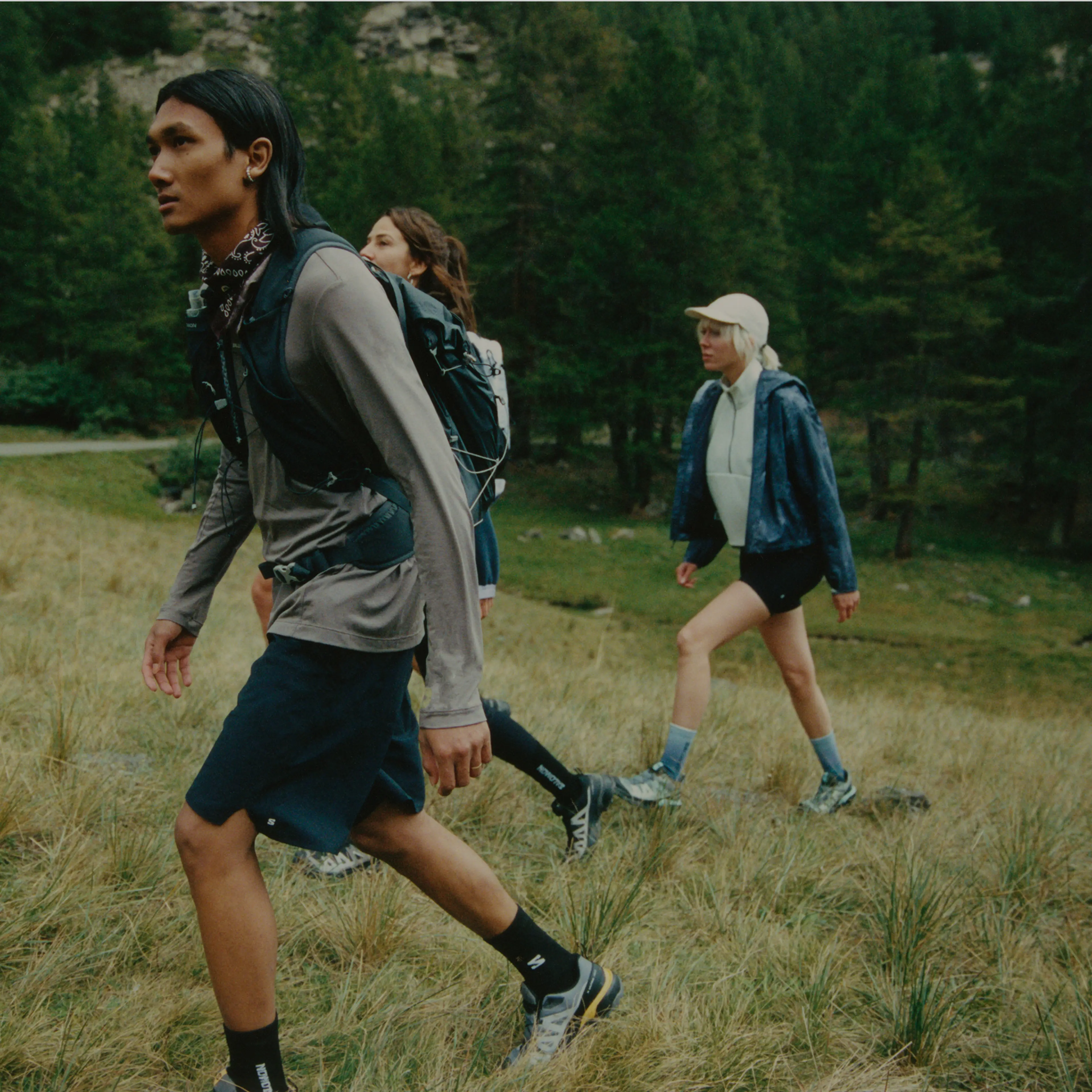 Three people hiking uphill through grassy terrain with backpacks, surrounded by a forested area.