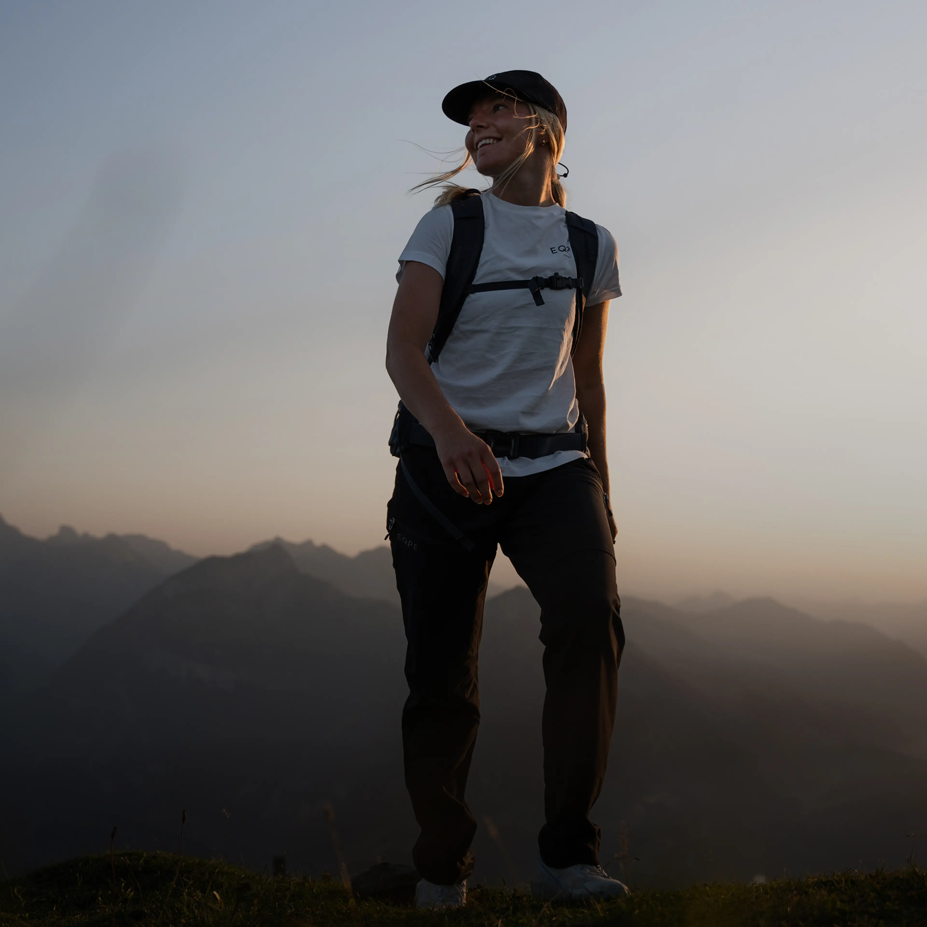 A person wearing a cap and backpack stands in front of a dusky sky, smiling and looking to the side, with mountain silhouettes in the background.