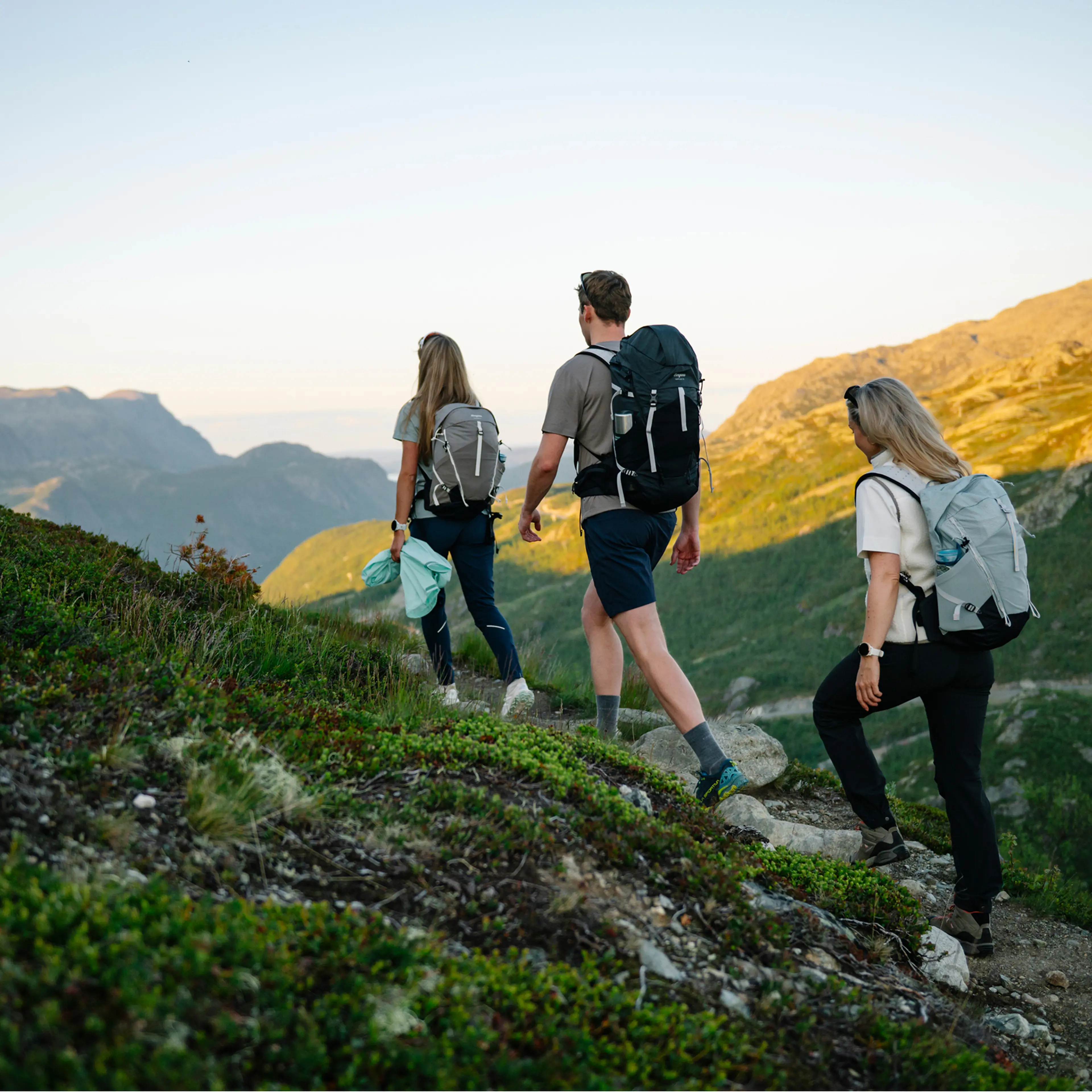 Three hikers with backpacks trekking up a grassy mountain path at sunset, overlooking distant peaks in the background.