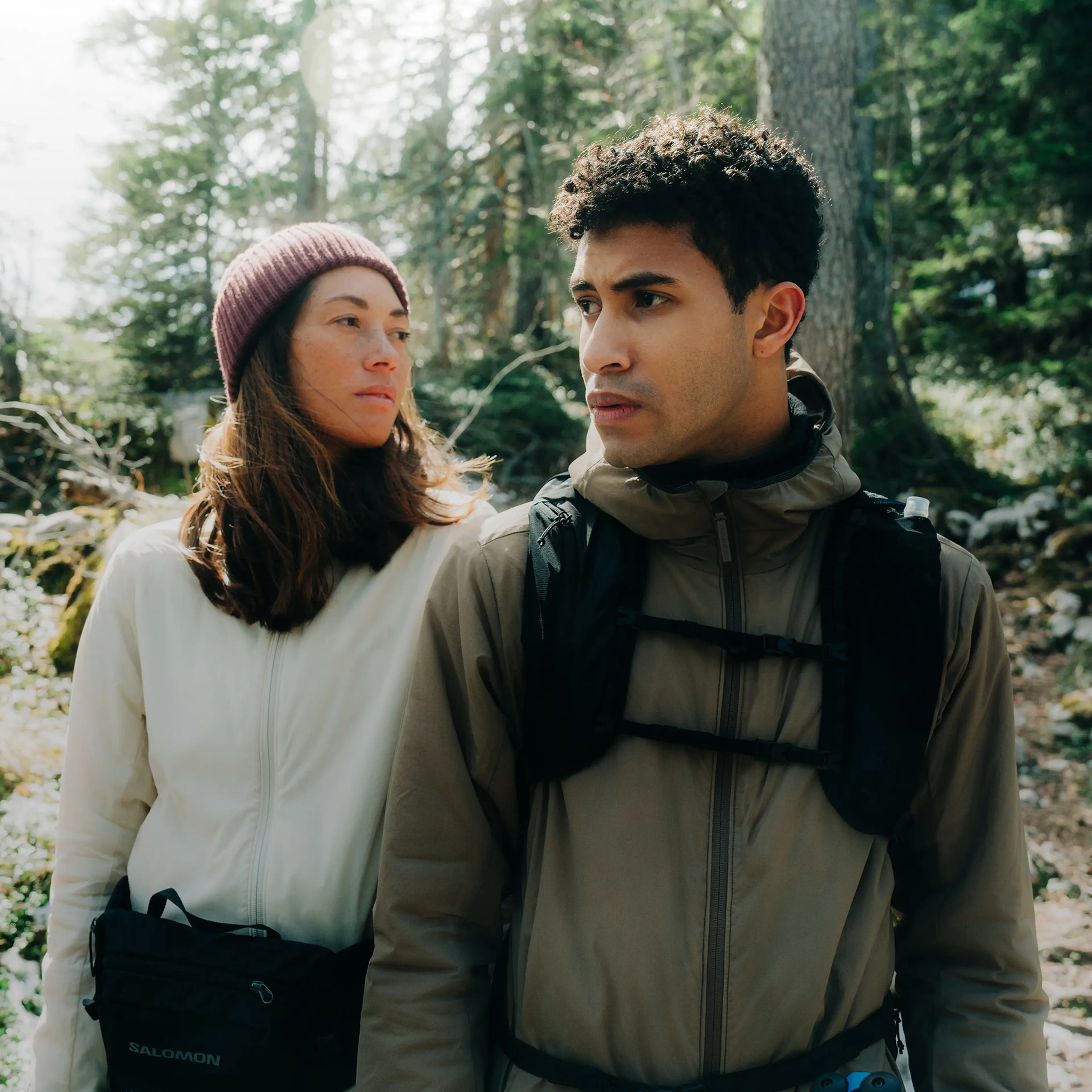 A man and a woman wearing outdoor gear stand in a forest, looking intently into the distance, surrounded by trees and sunlight.