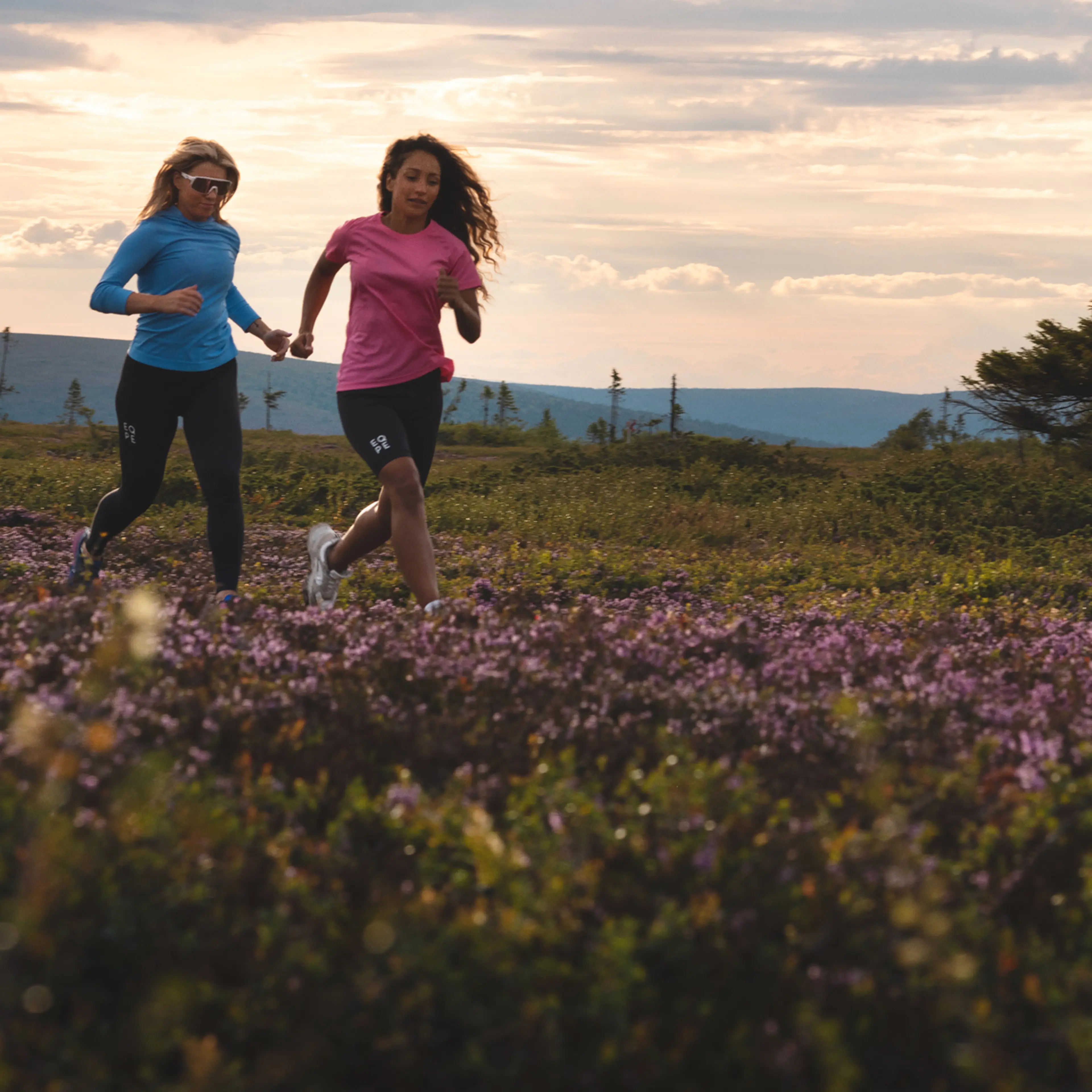 Two people jogging through a field of purple flowers with trees and hills in the background under a cloudy sky.